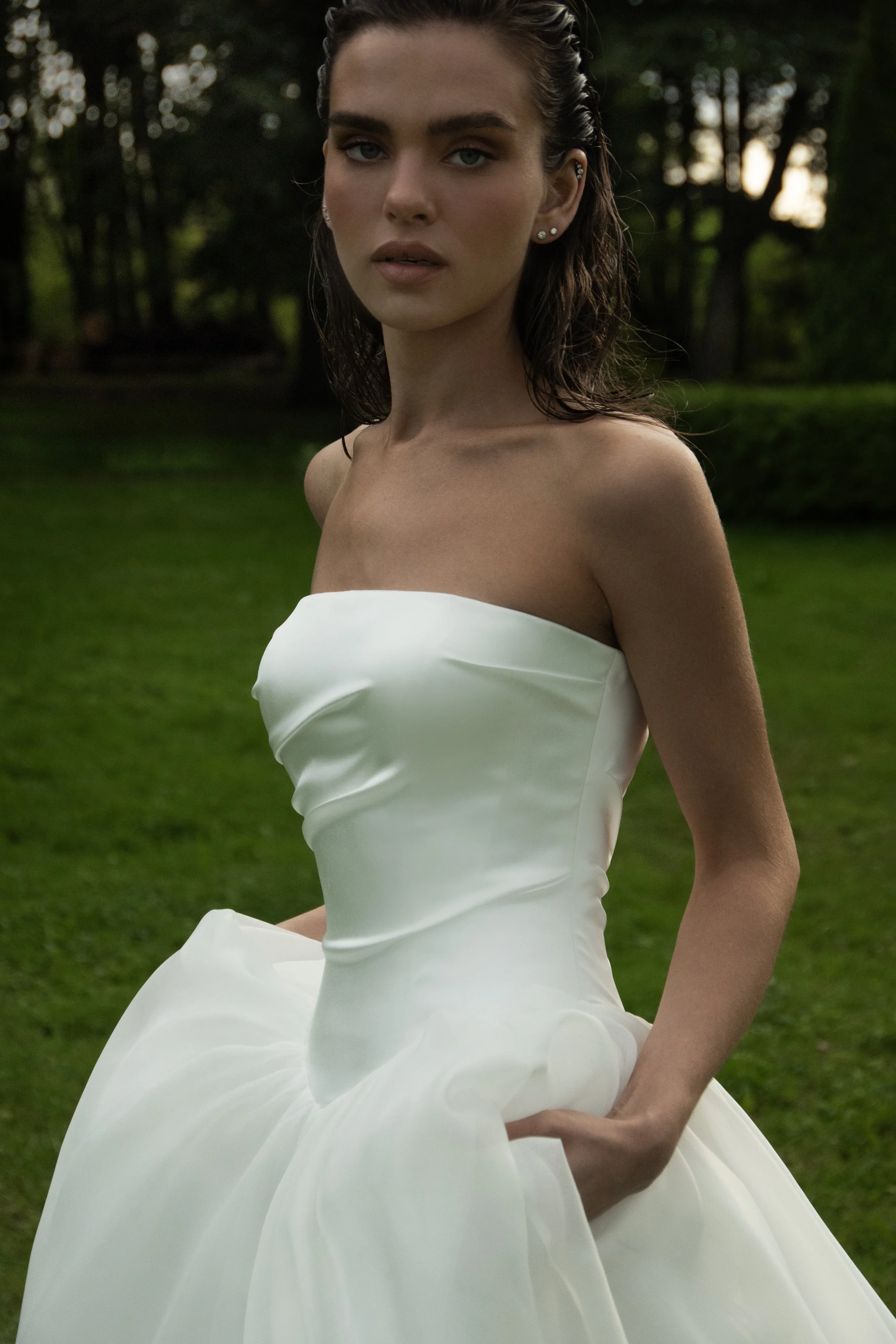 Detail shot of Solo wedding gown bodice by Romanova Atelier, crisp satin-like structure, bride holding floaty organza skirt, green lawn and trees in background.