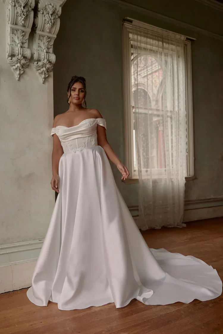 Brisbane bride wearing Alaska gown by Anna Campbell, off-the-shoulder wedding dress with floral embroidery and a sheer corset bodice, standing outdoors in front of a decorative stone staircase and wooden windows.