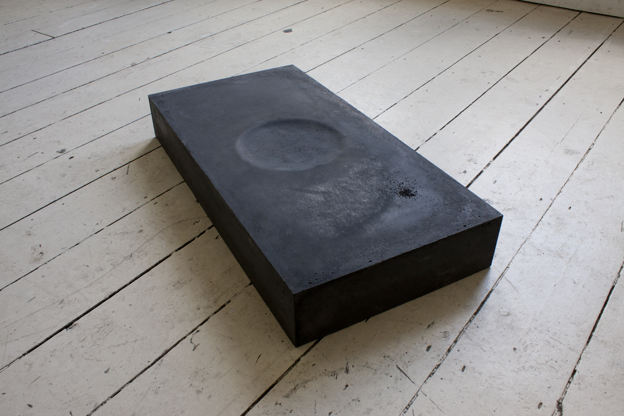 Hora Somni: for Vera Rubin, 2017 Cast black concrete with Papaver Somniferum seeds, 10 x 35 x 70cm Off to one corner of the work is a small pile of sleep-bringing poppy seeds. The piece is very heavy, bound by the laws of gravity. On its surface, it bears a shallow circular crater, the imprint of a disc-shaped galaxy or a head on a pillow. While others are sleeping, astronomers override their circadian rhythms to look for points of light in a sea of blackness.