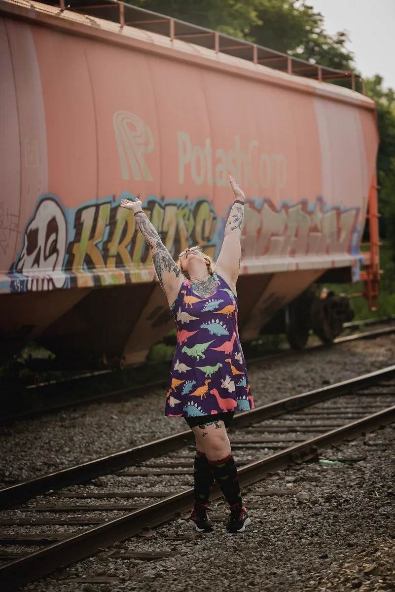 A woman, Kelly Mendenhall, wearing a multi-colored dress adorned with cartoon dinosaurs, rejoices with her hands in the air, with a train car in the background