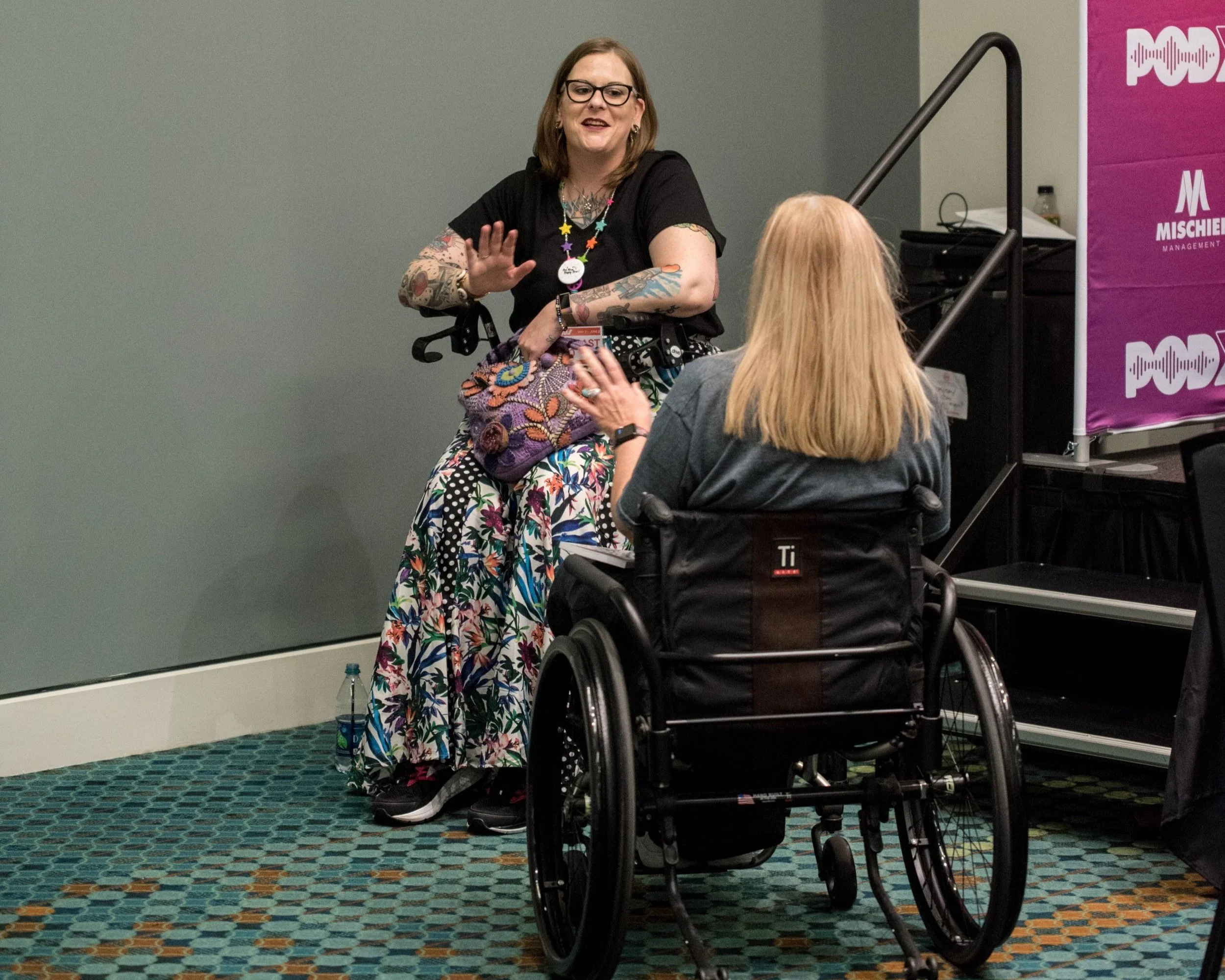 Image of two women talking. One is sitting in a wheelchair with her back to the camera, the other is sitting on her Rollator walker, facing the camera. The two women are engrossed in conversation.