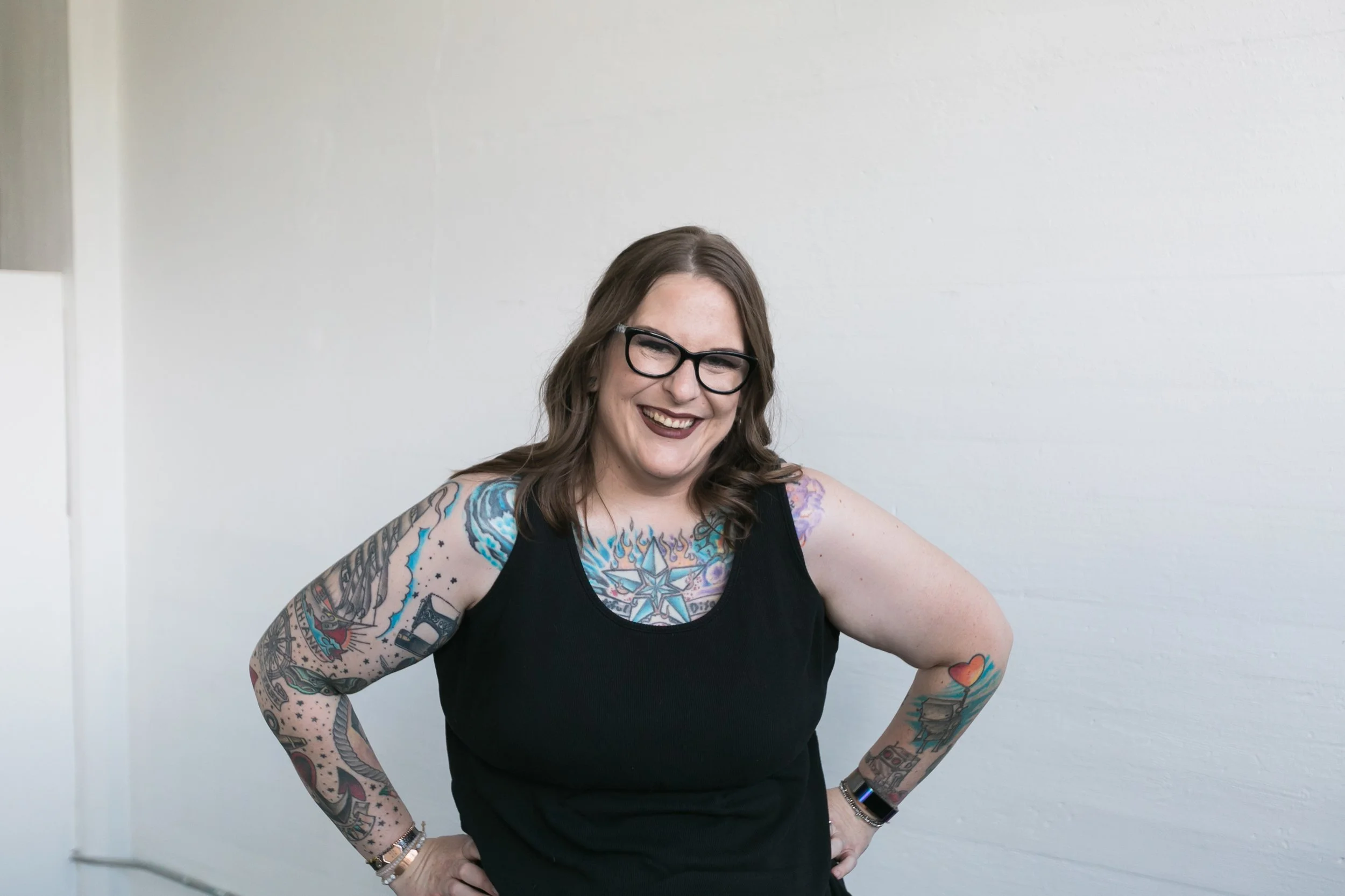 The image of a heavily tattooed woman, smiling, standing in front of a white wall. She is wearing a tank top and glasses and appears to be laughing.
