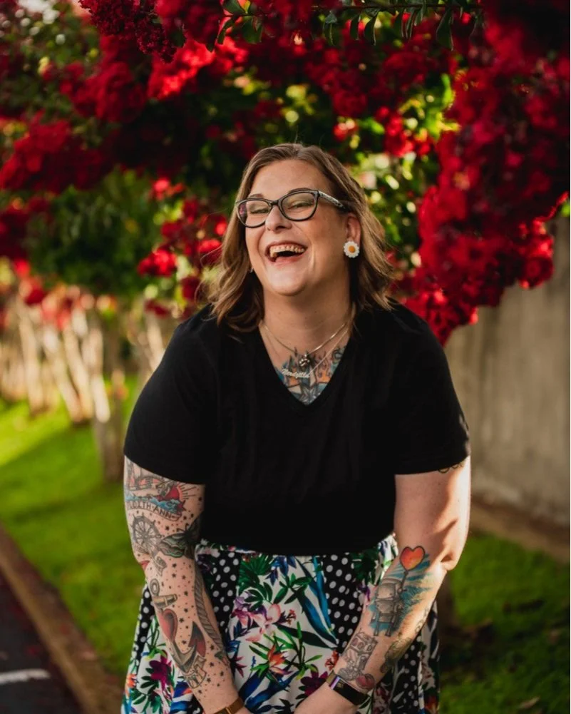 An image of a woman smiling, wearing a flora skirt with black top. Red flowers and green grass are behind her. She has a myriad of tattoos on her arms and chest.