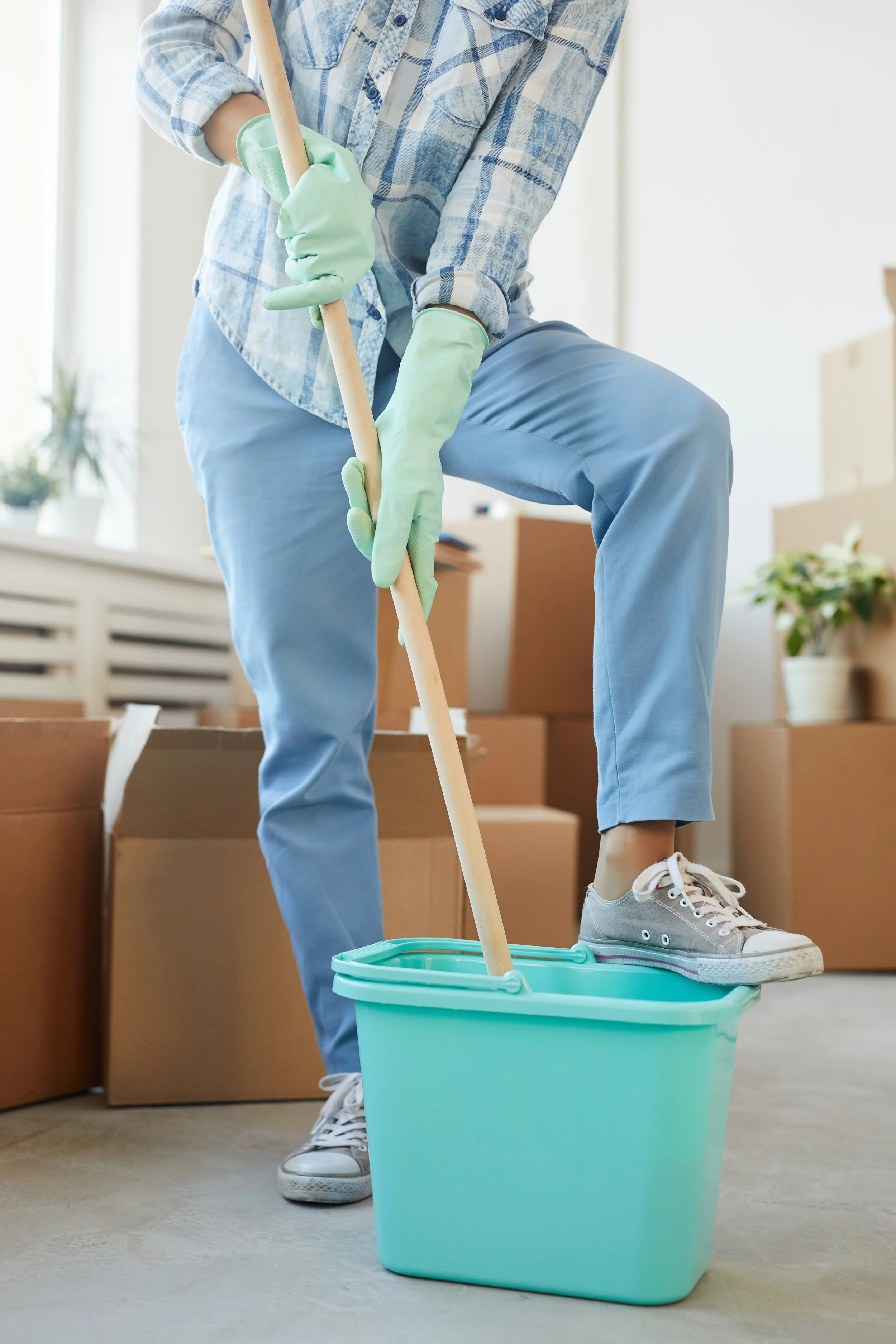 Person wearing gloves and casual clothes mopping the floor in a moving or storage room with cardboard boxes.