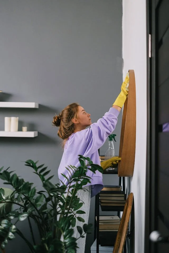 A woman wearing yellow gloves cleaning a wooden shelf on a wall in a modern living space with gray walls and white floating shelves.