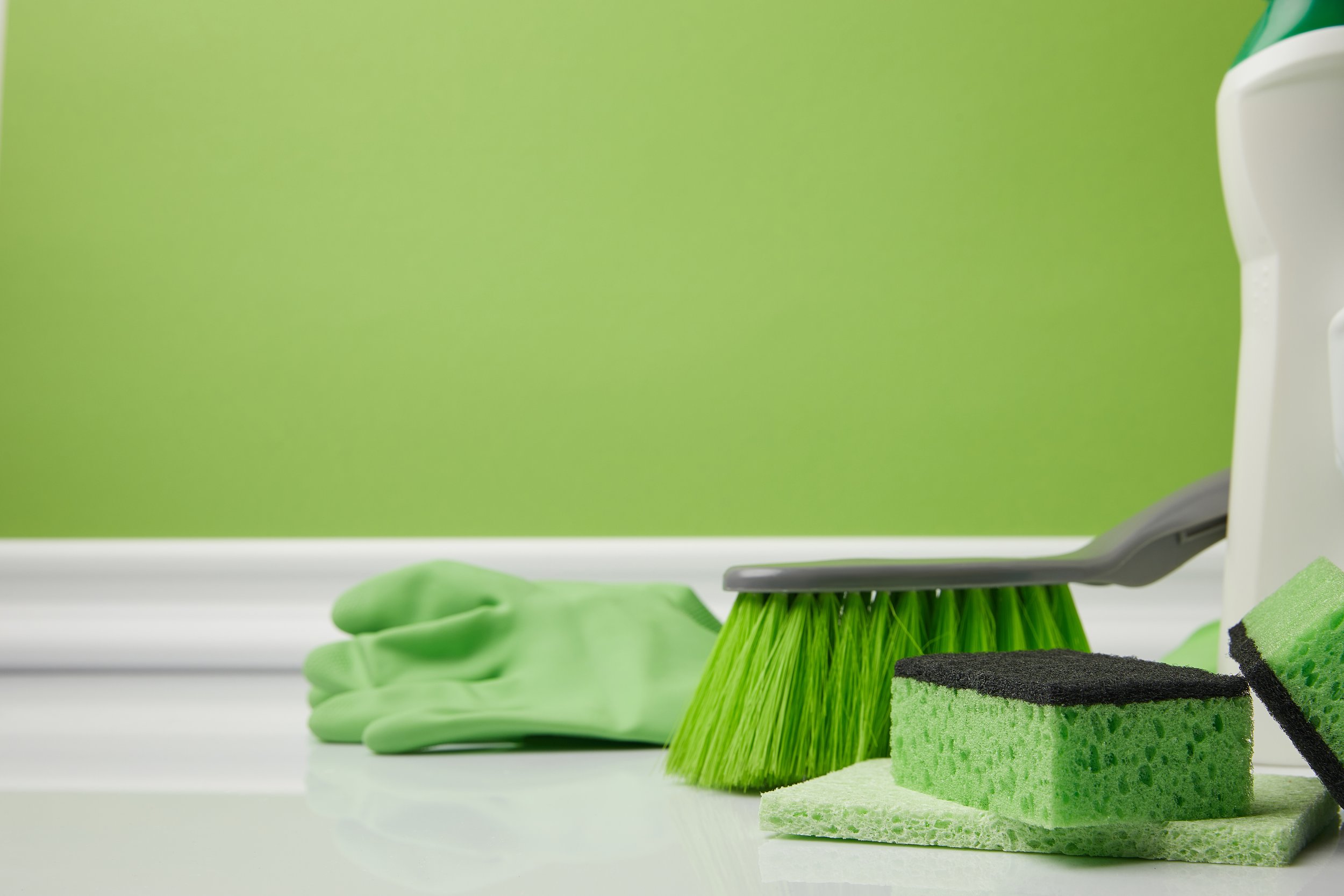 Green cleaning gloves, a green dishbrush, and green cleaning sponges on a white surface with a light green wall in the background.