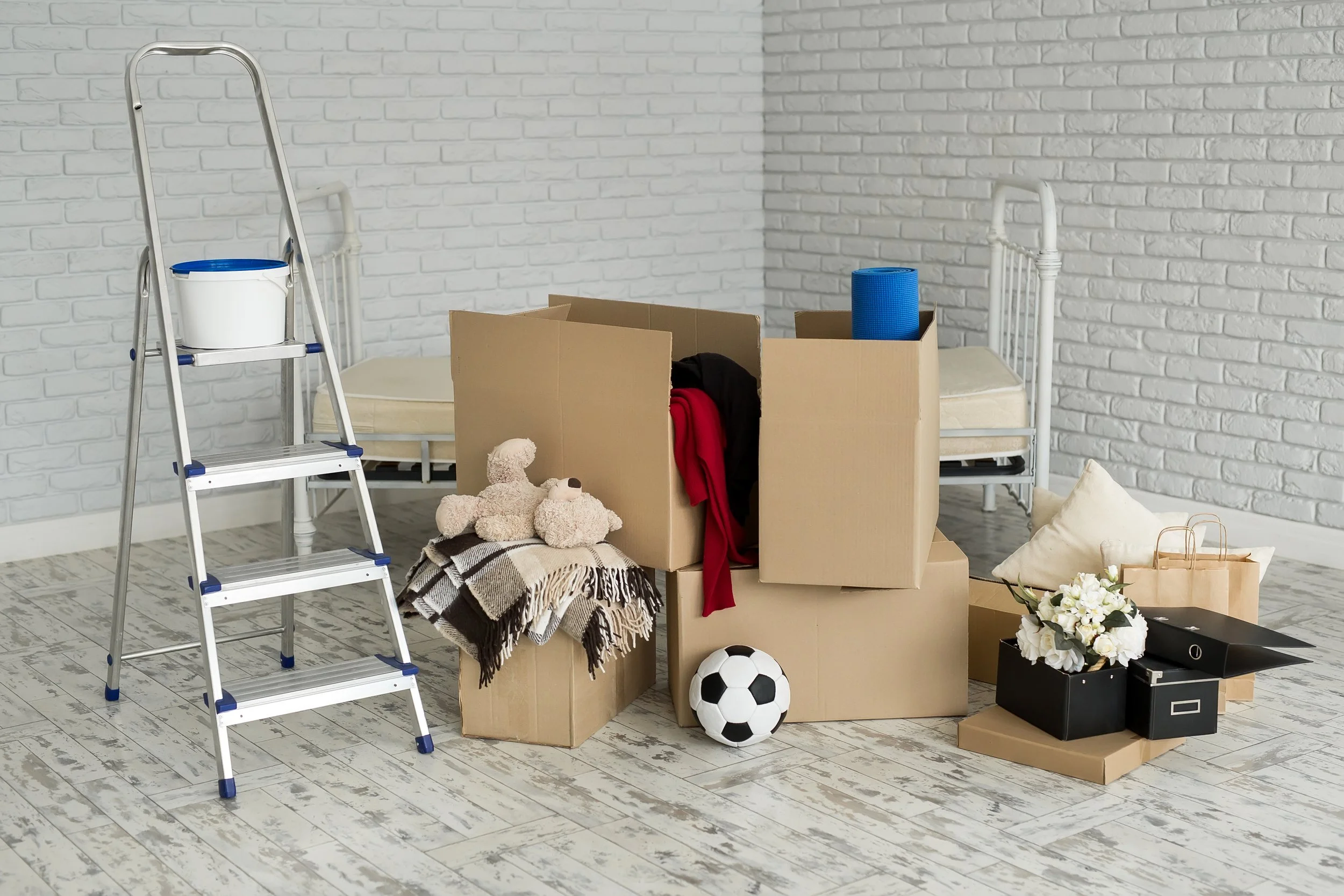 Room filled with moving boxes, a small ladder with a bucket, toys, folded blankets, a soccer ball, flowers, and cushions, against white brick walls and wooden floors.