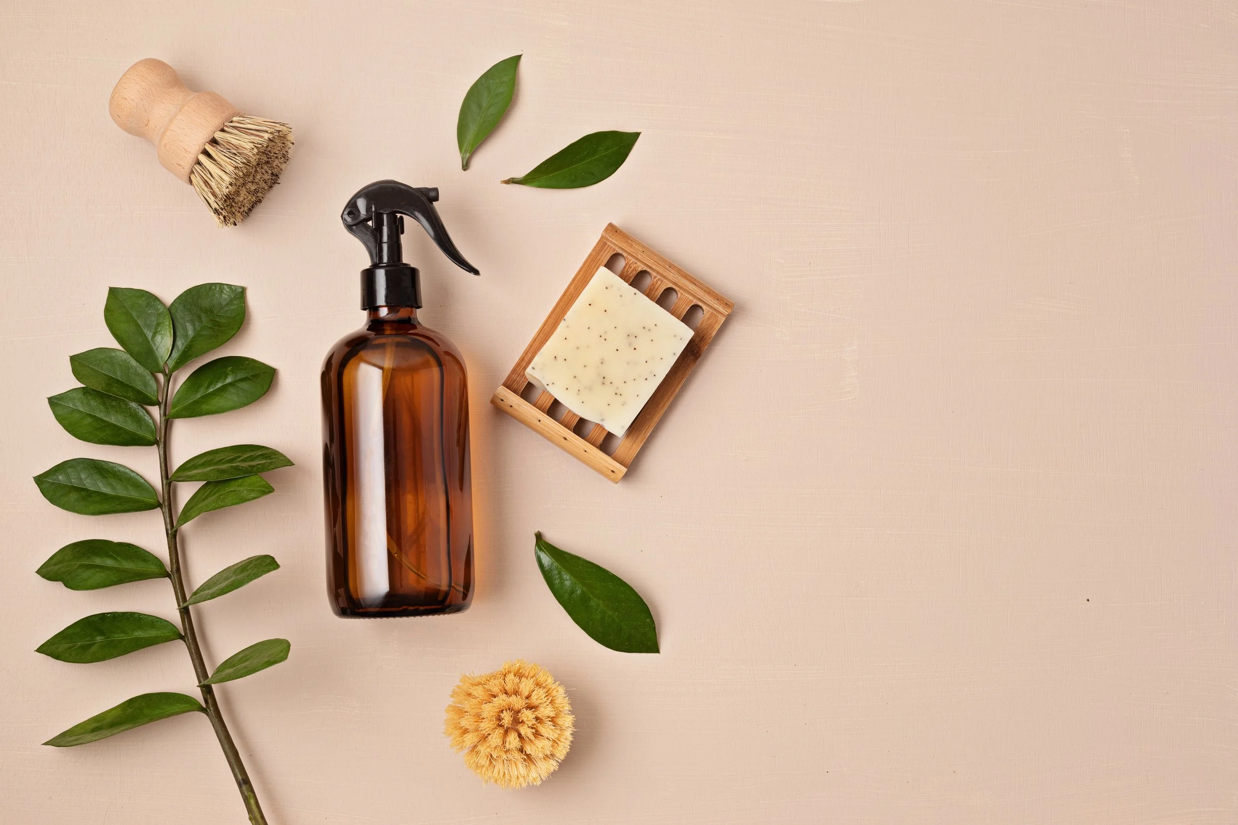 Glass spray bottle, natural soap bar on bamboo soap dish, sponge brush, green leaves, and a yellow sponge on a beige background, arranged for eco-friendly cleaning or skincare.