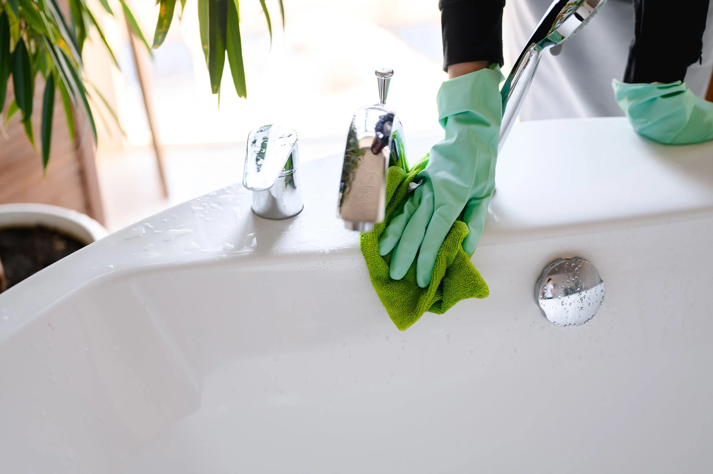 Person cleaning a white bathroom sink with a green cloth, wearing green rubber gloves.