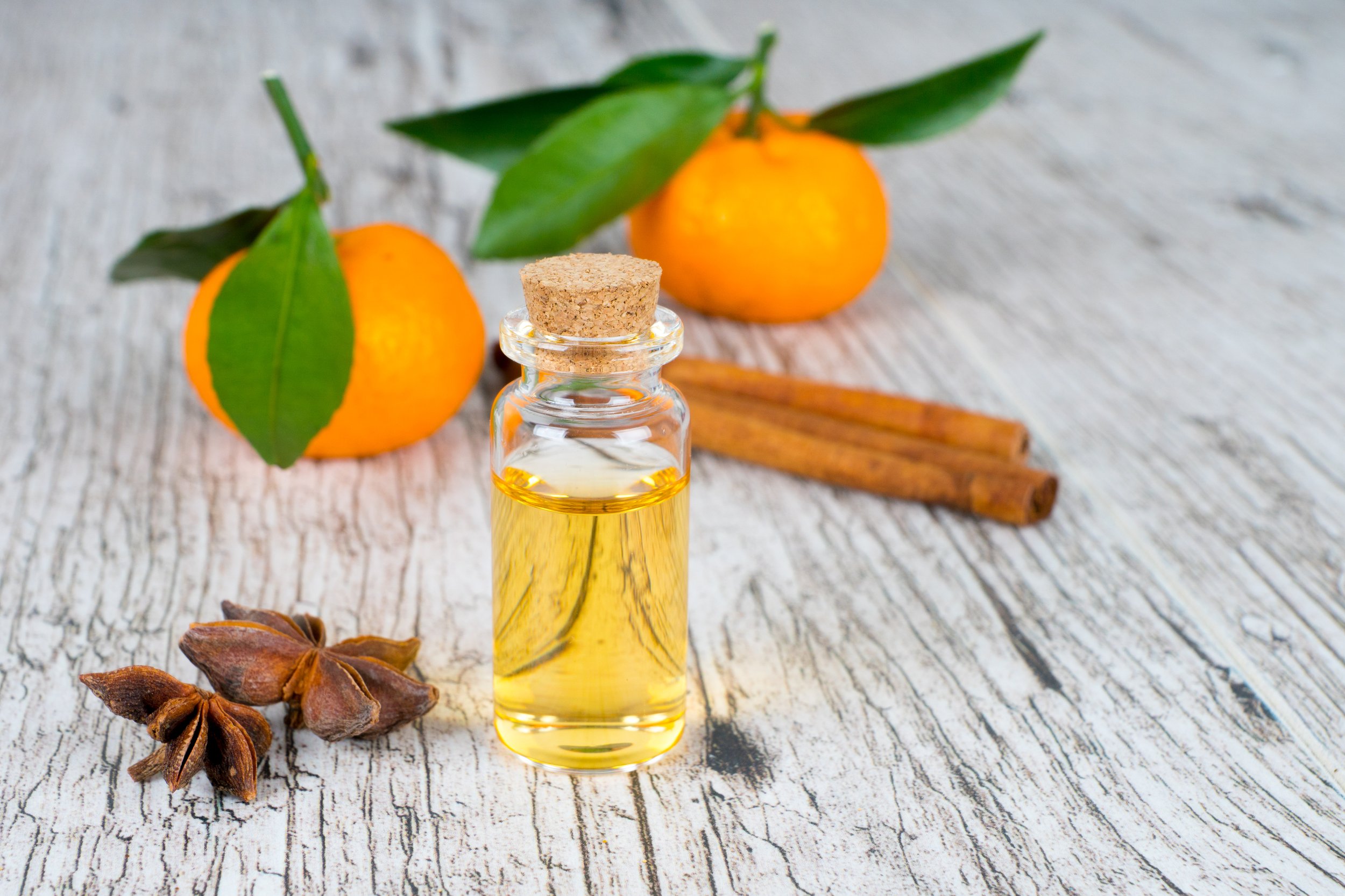 A small glass bottle of yellow oil with a cork stopper, two mandarins with green leaves, two cinnamon sticks, and star anise on a rustic wooden surface.