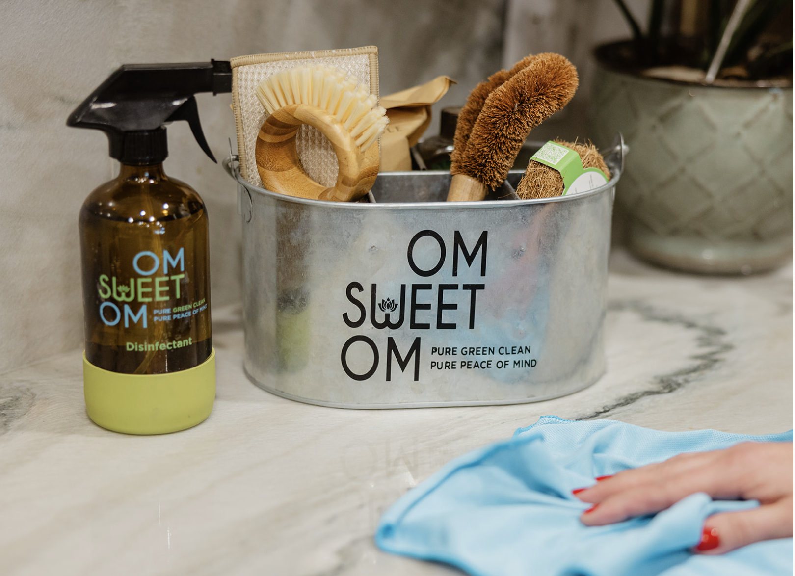 A metal container with the words 'OM Sweet OM' and the phrase 'Pure Green Clean, Pure Peace of Mind' printed on it, holding cleaning brushes and a disinfectant spray bottle labeled 'OM Sweet OM'. A person's hand with red painted nails is wiping a surface with a blue cloth in the foreground.