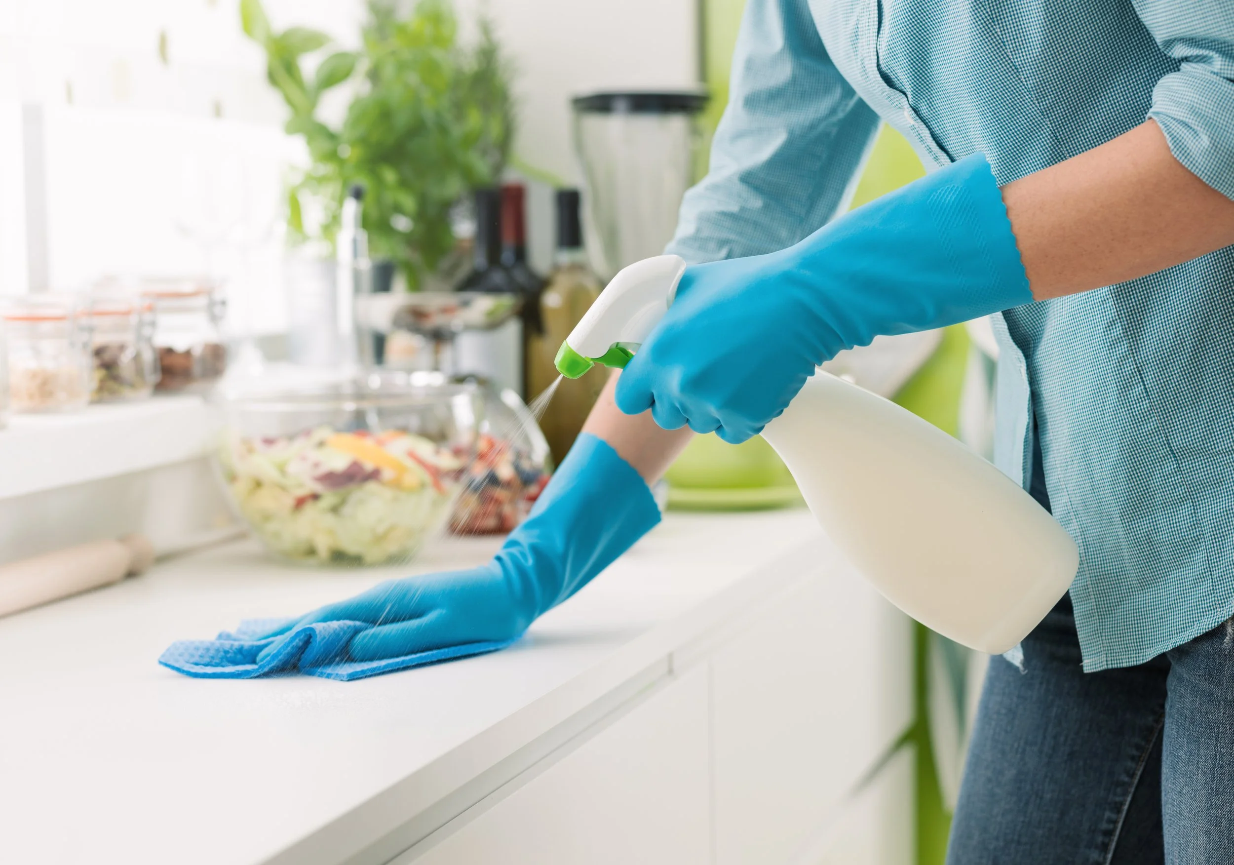 Person wearing blue gloves cleaning a kitchen counter with spray bottle and cloth, with a bowl of salad and kitchen appliances in the background.