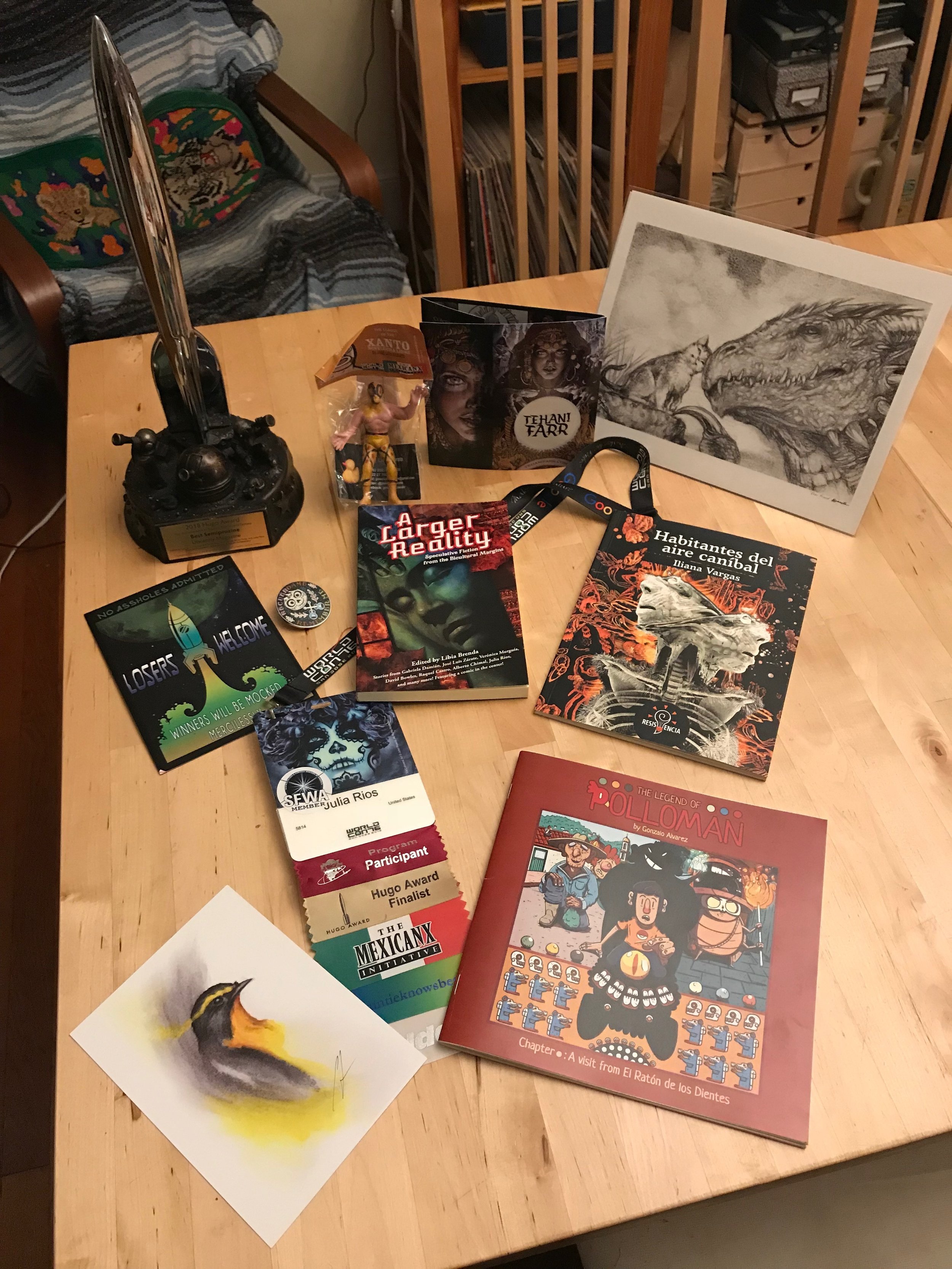 A haul of Mexicanx Initiative works and Worldcon 76 mementos Julia Rios cherishes. Back left to right: Hugo Award with base designed by Sara Felix and Vincent Villafranca, lucha libre figure from JosĂ© Luis Zárate, art of Tehani Farr, kitty and drago print by DIanita CerĂłn. Middle left to right: Invitation to the Hugo Losers Party, *Coco* enamel pin, a copy of *A Larger Reality*, *Habitantes del aire cannĂbal* by Iliana Vargas. Bottom left to right: Bird art print by Cody Jimenez, Julia Rios's Worldcon 76 name badge with art by John Picacio and a Mexicanx Initiative ribbon, *The Legend of Polloman* by Gonzalo Alvarez. (Photo by Julia Rios)
