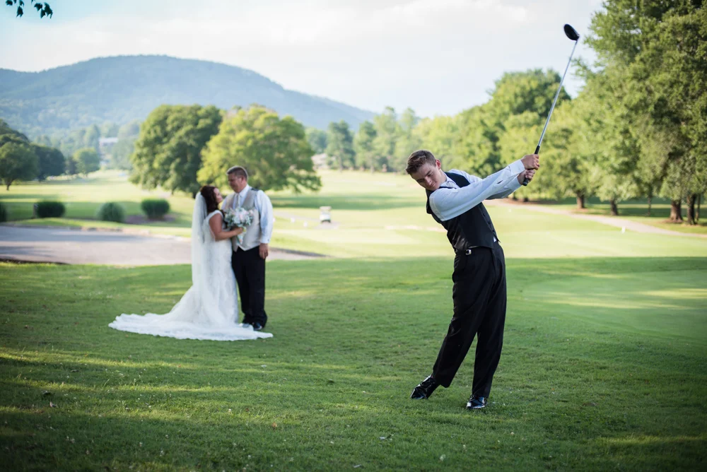 Bride and groom looking at each other in the background while son swings golf club on a golf course in the foreground