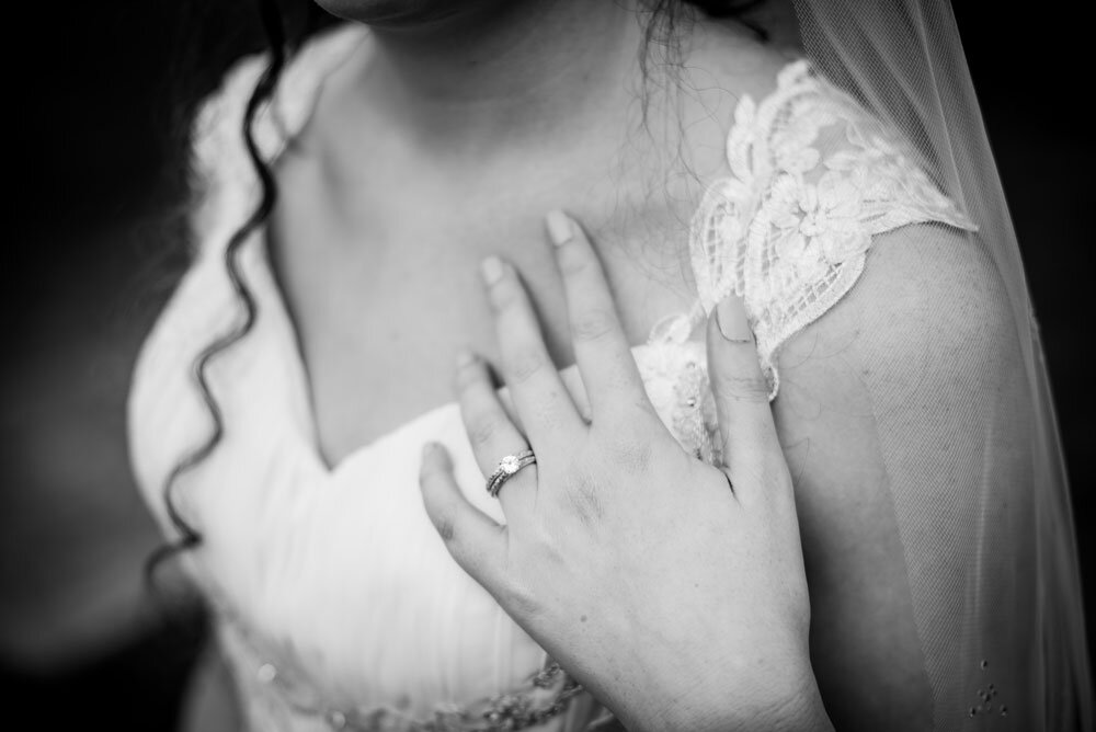 Bride holding her left hand to her shoulder to display her wedding ring next to her wedding gown.