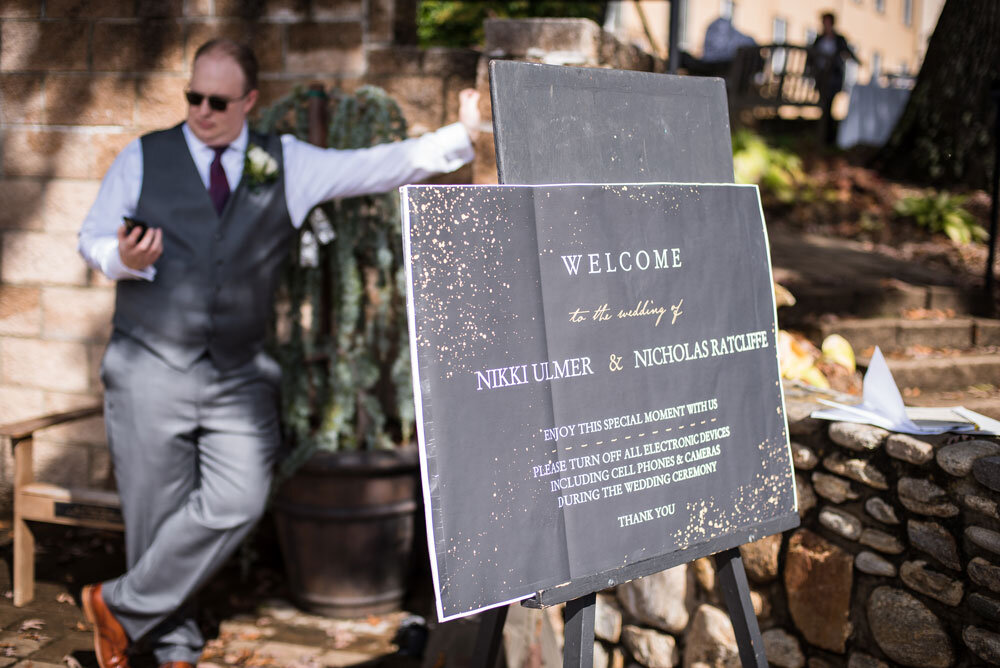 Groom holding cell phone behind a sign at the entrance of his wedding asking guests to turn their cell phones off