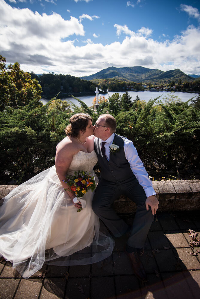 Bride and groom sitting on a ledge kissing with Lake Junaluska in the background.