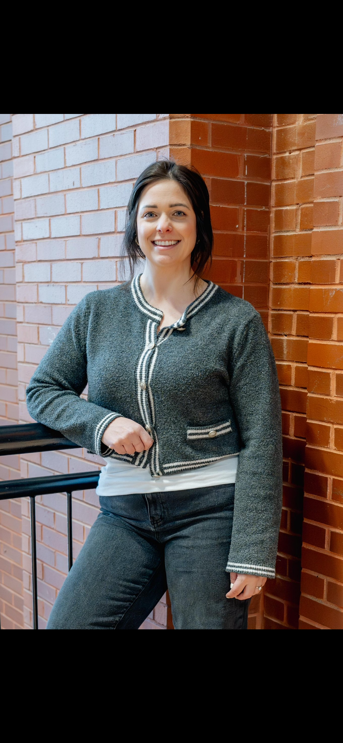 A woman standing outdoors near a brick wall, smiling, wearing a grey jacket with white and black trim, a white shirt underneath, and black jeans.