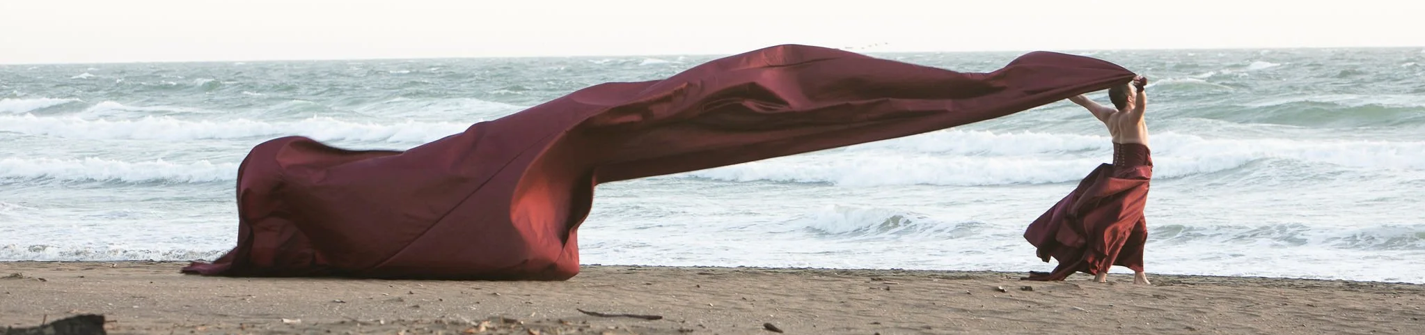 A dancer stands at the shoreline holding a long piece of flowing fabric above his head.