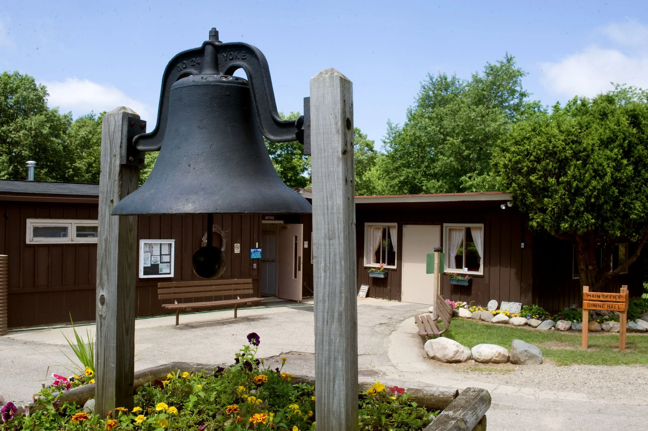 large cast iron bell sits in foreground, in front of single-level building. There are wooden benches, white doors and windows, and a wooden sign that says Main Office and Dining Hall