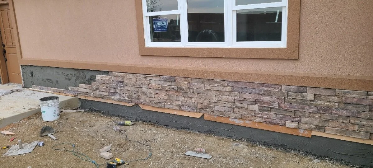 Construction site with partial stone veneer installation on building exterior, white window above, tools and materials on the ground.