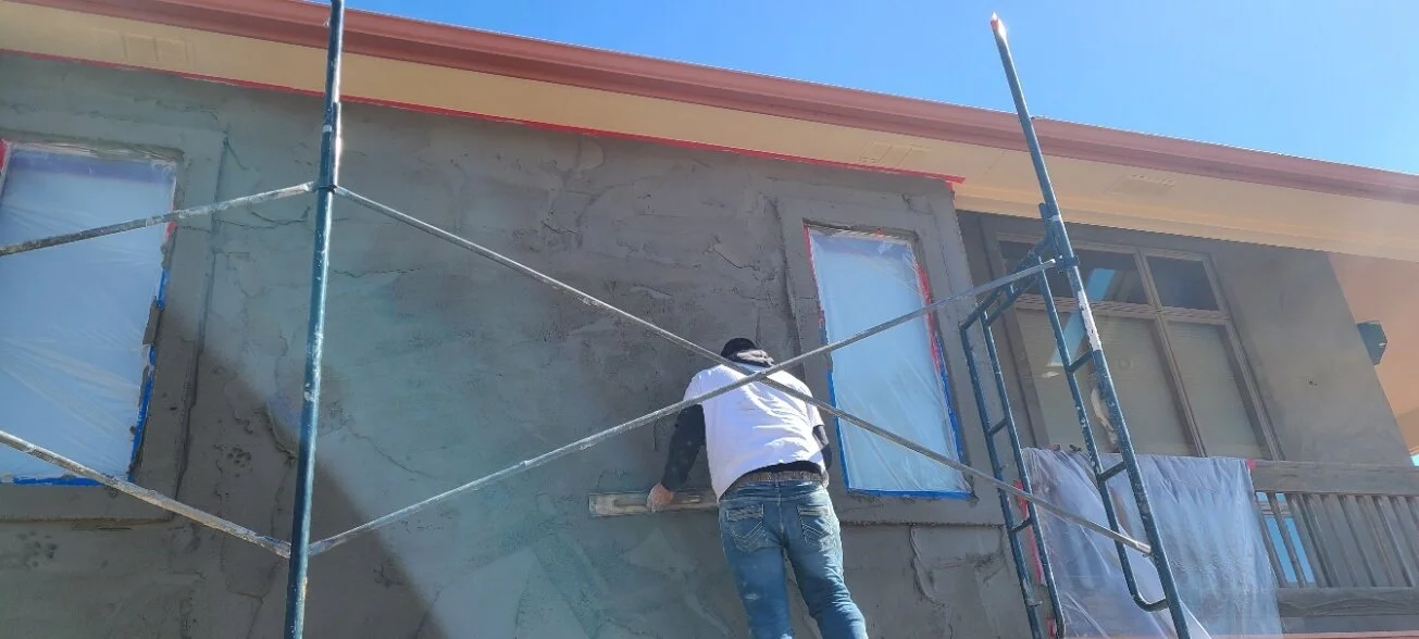 Worker applying stucco on a house exterior using a trowel, scaffolding present, windows covered for protection.
