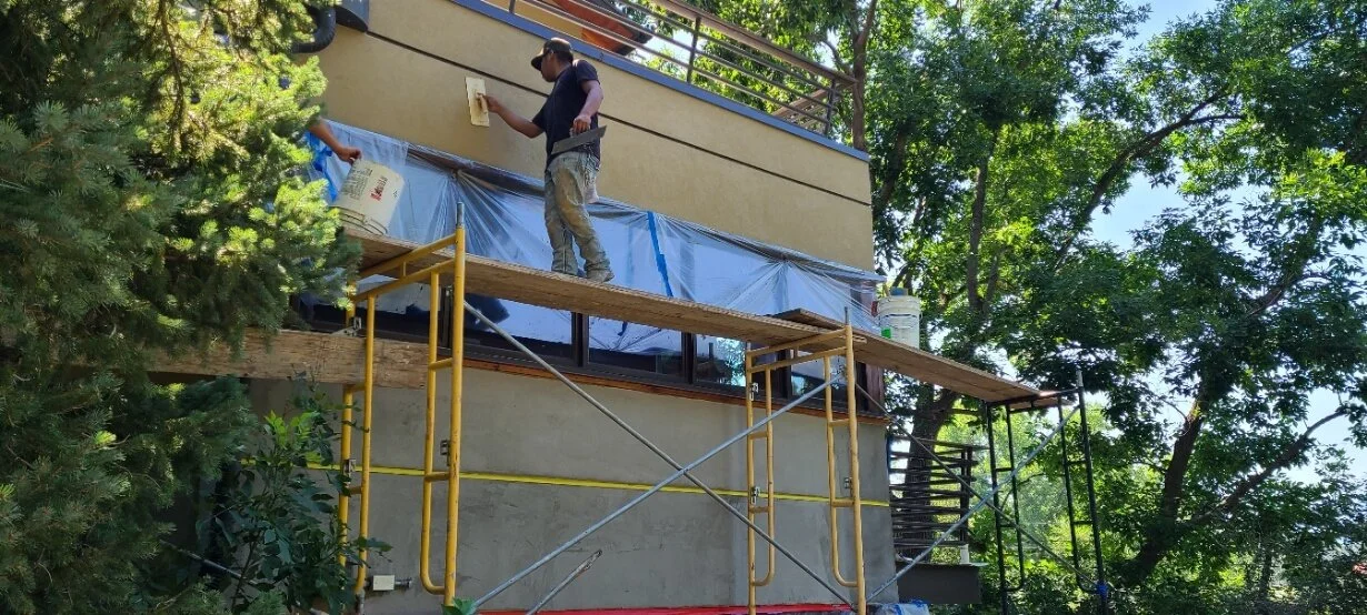 Construction workers applying stucco to the exterior of a house, standing on scaffolding, surrounded by trees.