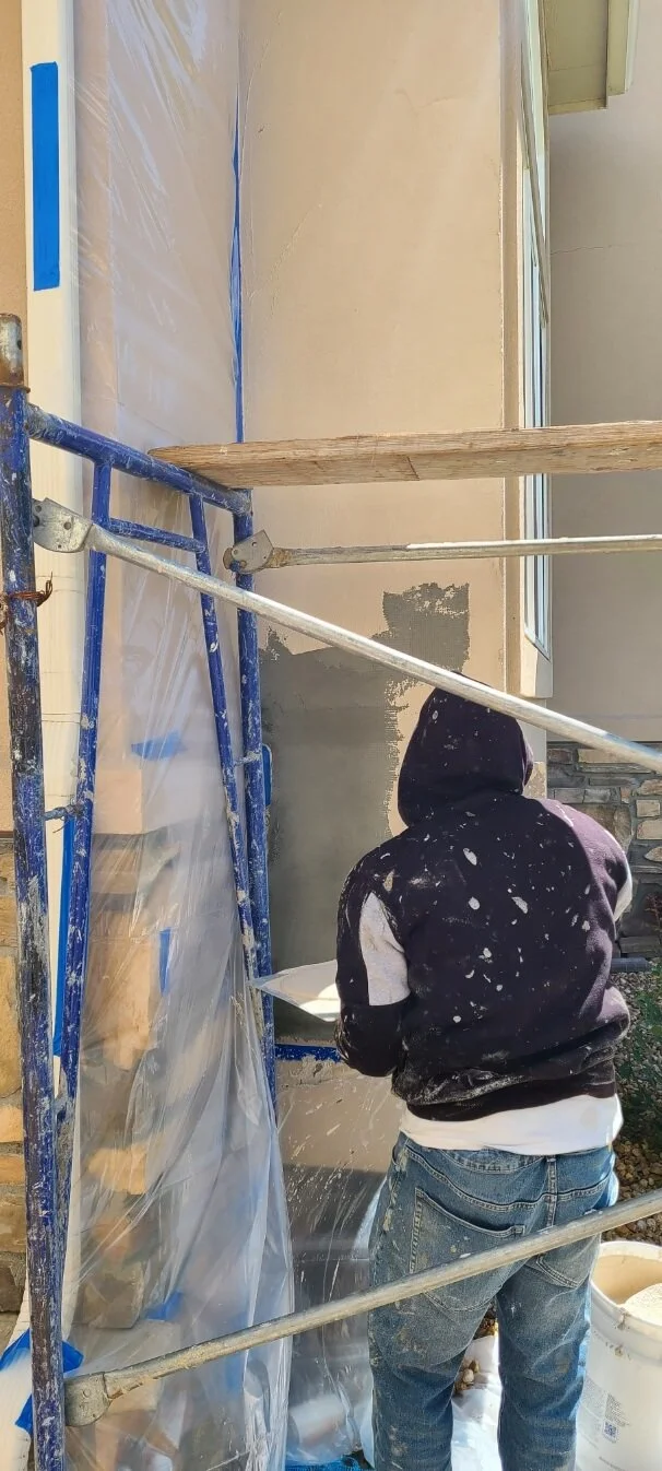 Person in hoodie applying plaster on wall, standing on scaffolding