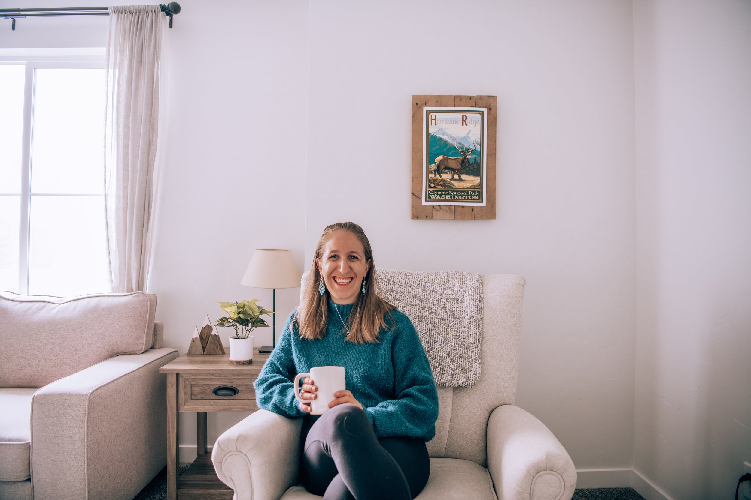 A woman sitting on a beige armchair in a living room, holding a white mug and smiling. There is a beige sofa, a side table with a plant and decorative items, and a window with light-colored curtains. A framed poster of Hurricane Ridge at Olympic National Park in Washington is hanging on the wall behind her.