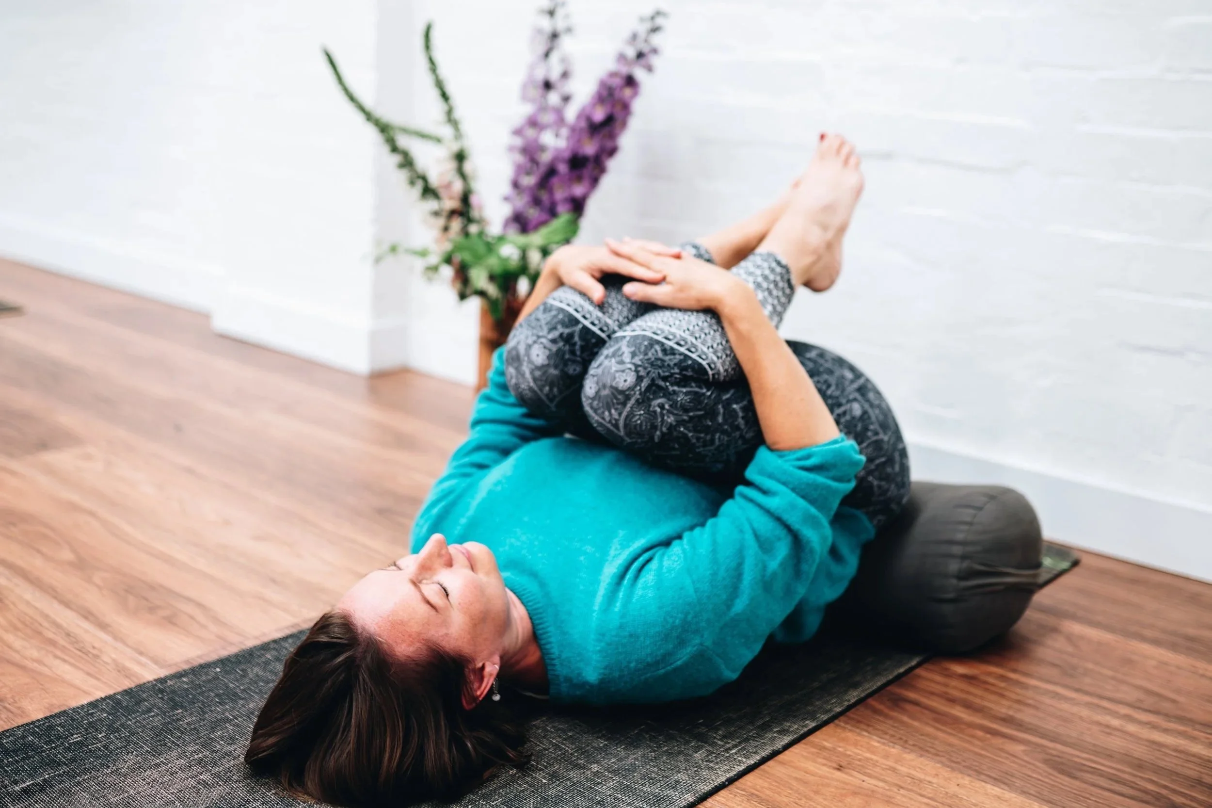 A woman practicing yoga together on a black mat inside a room with wooden floor and white wall. The woman is lying on her back with knees bent and arms crossed over her stomach, with a relaxed expression.