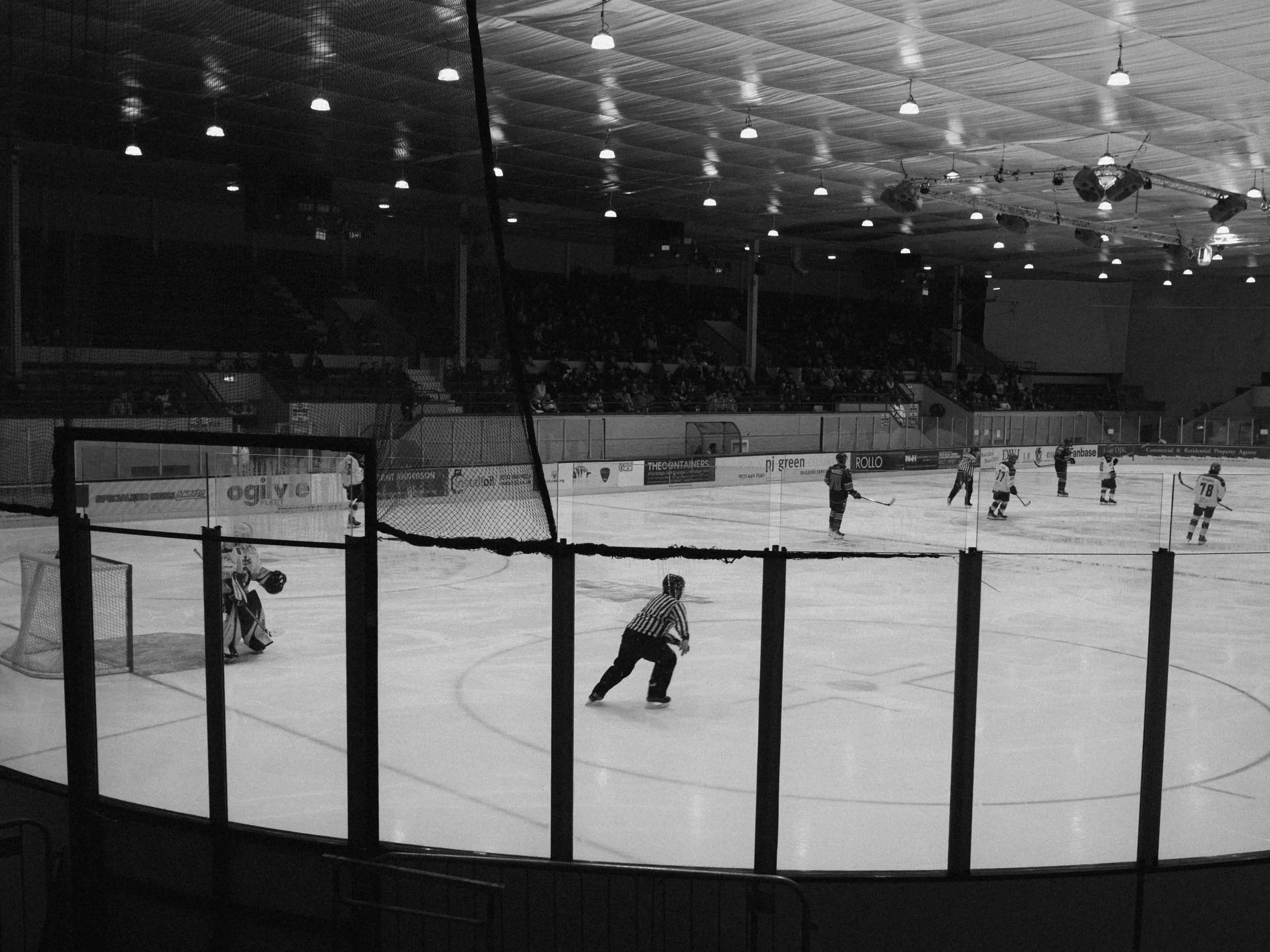 An indoor ice hockey game with players on the ice, a goalie in front of the net, a referee skating near the goal, and spectators watching from the stands.