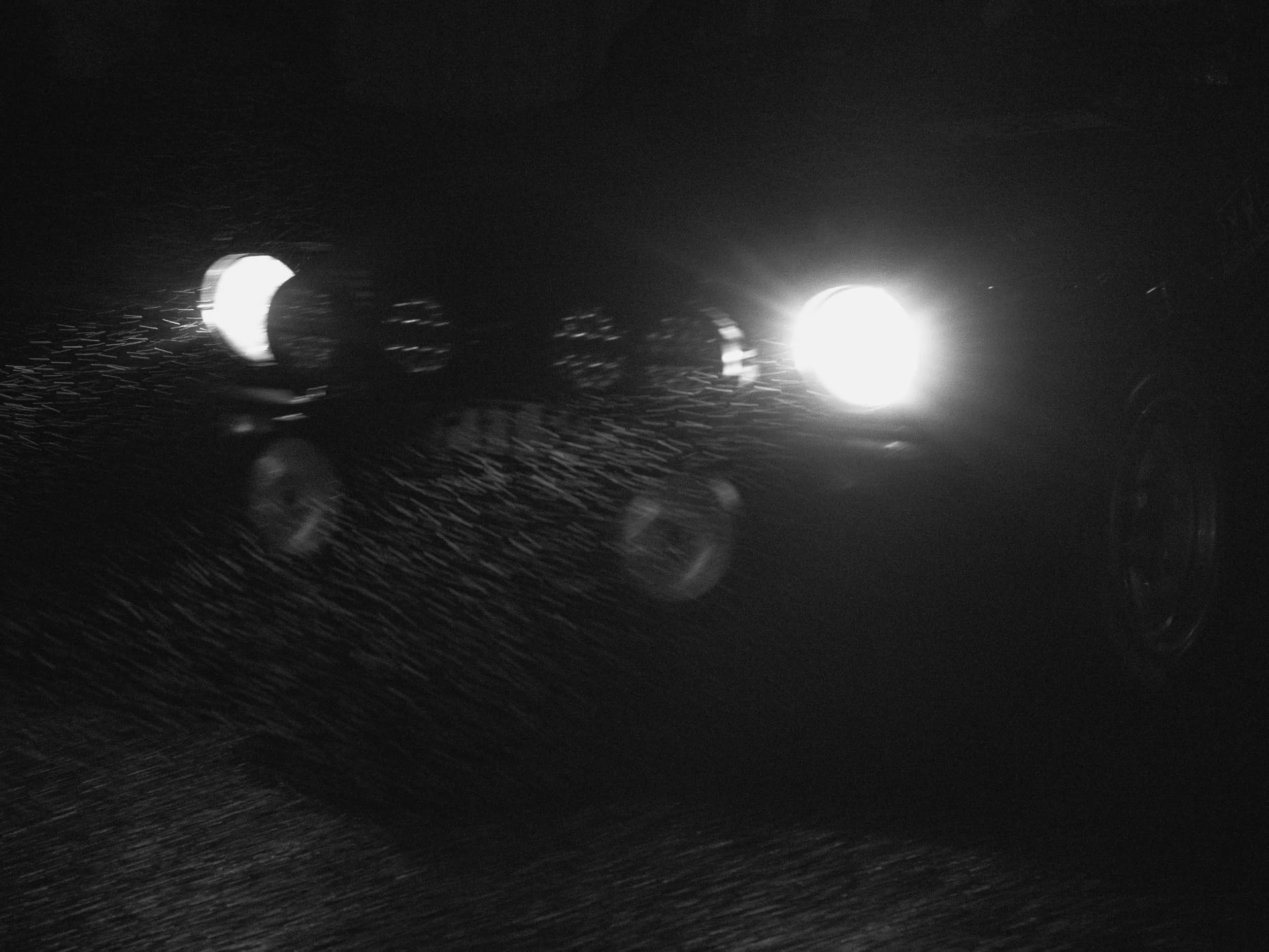A black and white photo of a car's front, with headlights on, driving through rain at night.
