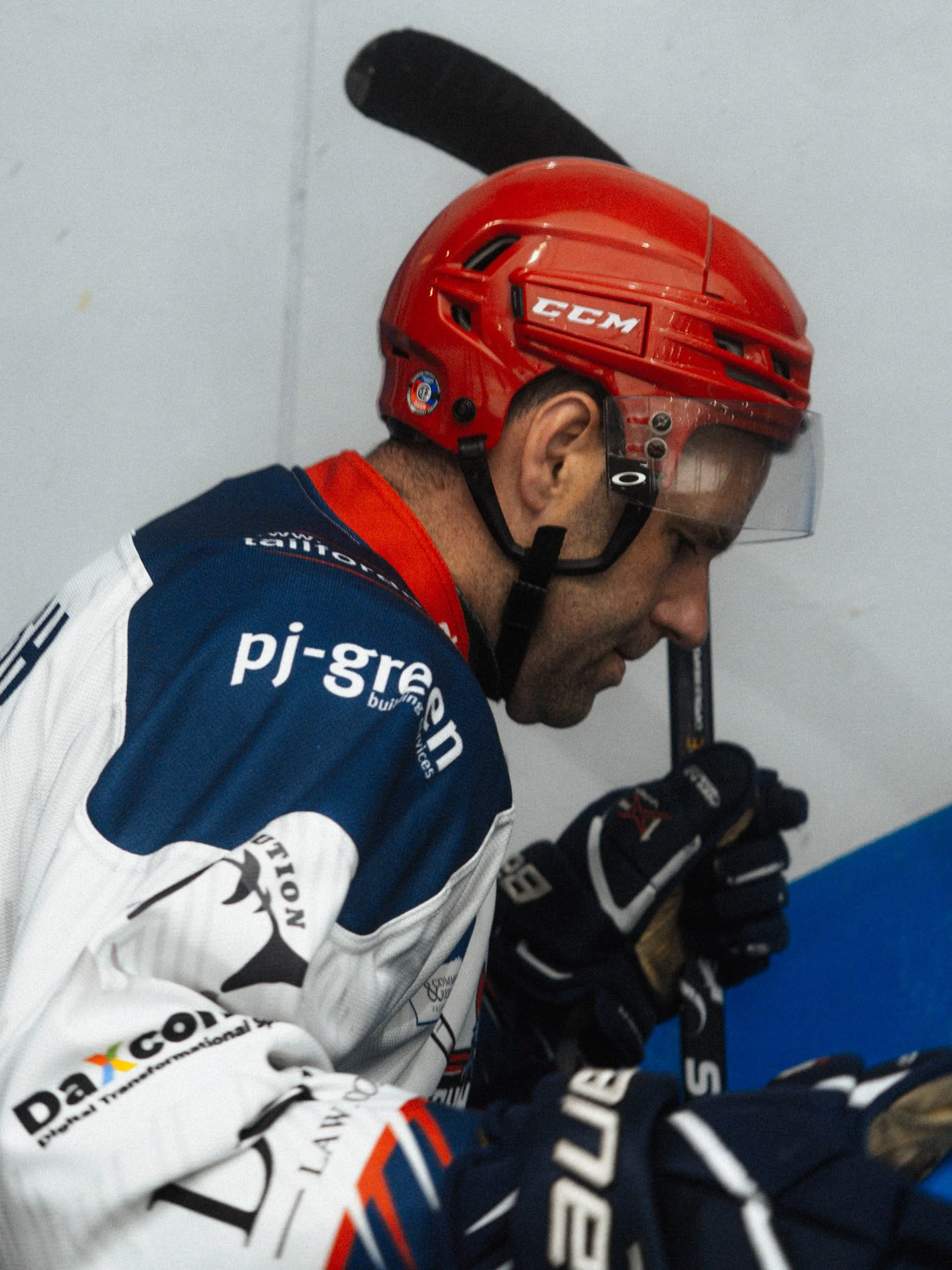 A male ice hockey player in a red helmet and blue and white jersey with sponsors, holding a hockey stick and looking down in concentration.