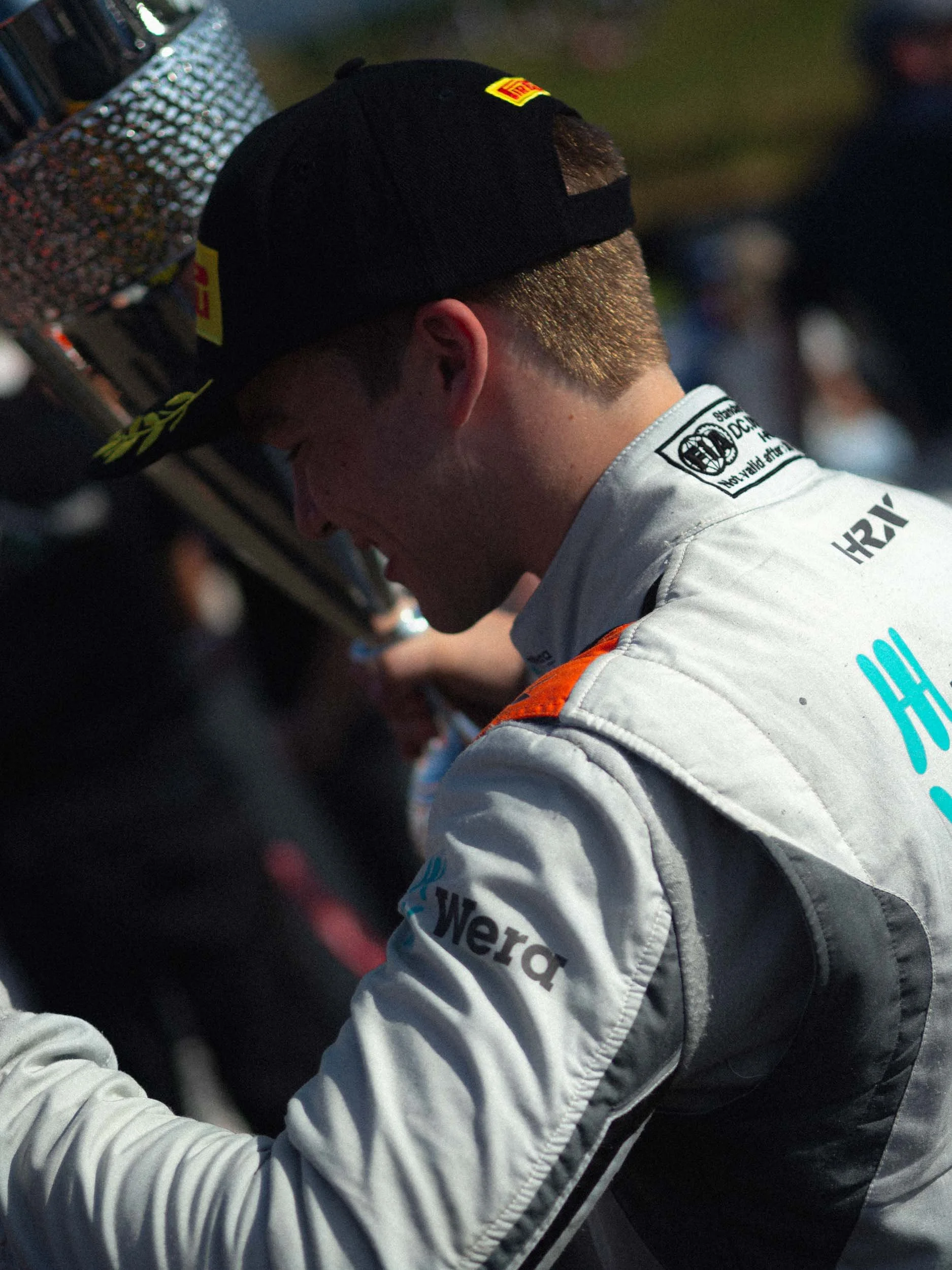 A male race car driver in a white racing suit with sponsorships, wearing a black cap with a yellow logo, with people in the background.
