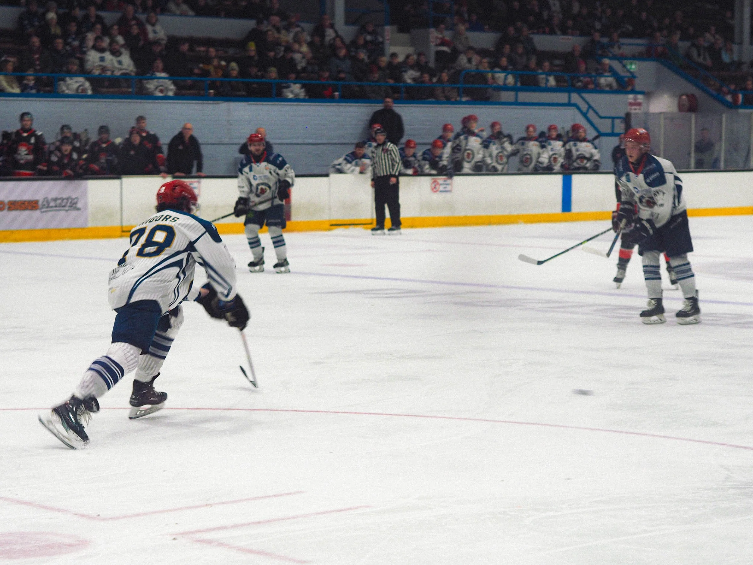 Hockey players on ice rink during game, with fans in stands and players on bench in background.