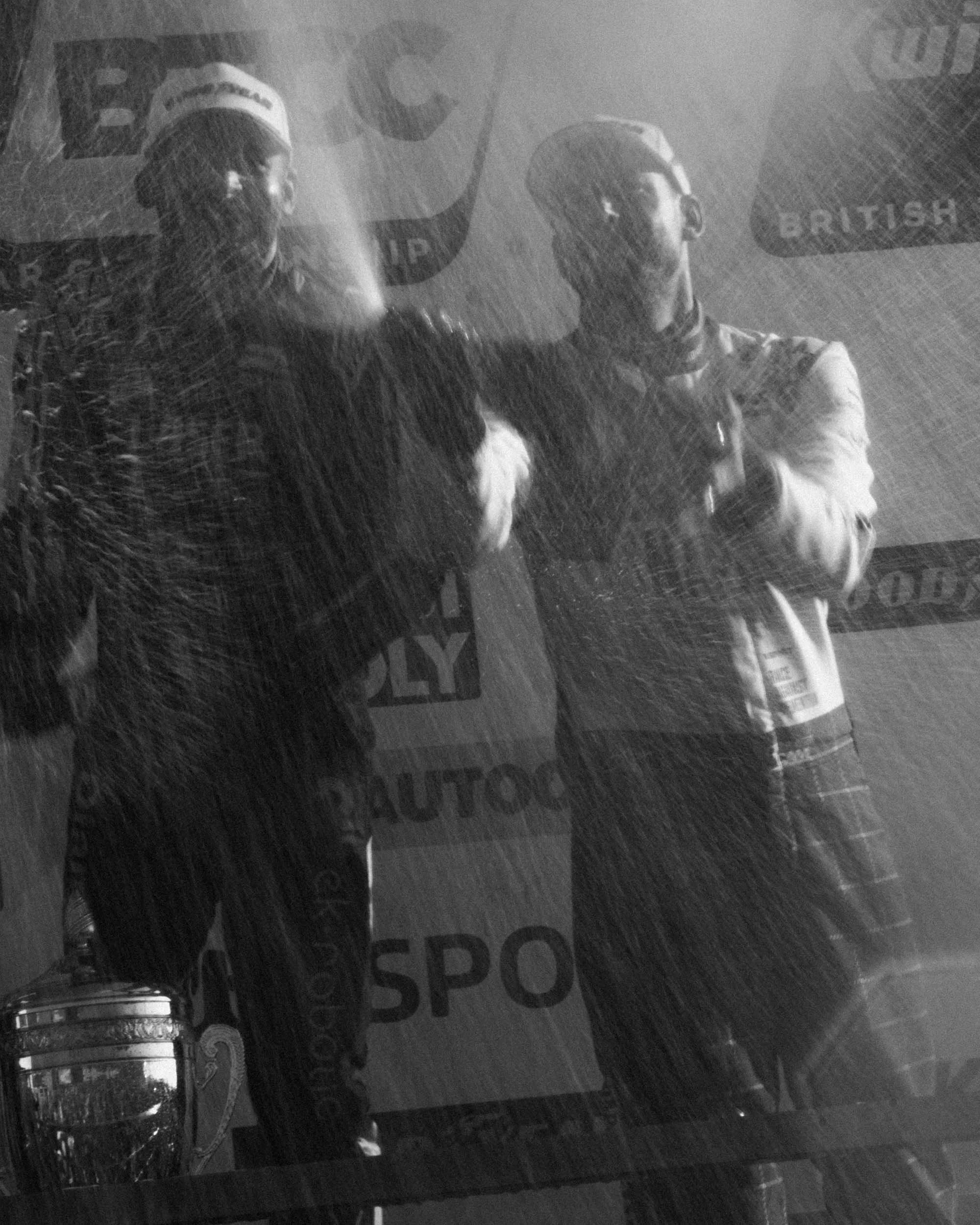 Two race car drivers in racing suits stood on the winners podium, champagne is actively being sprayed, with a trophy and promotional banners in the background.
