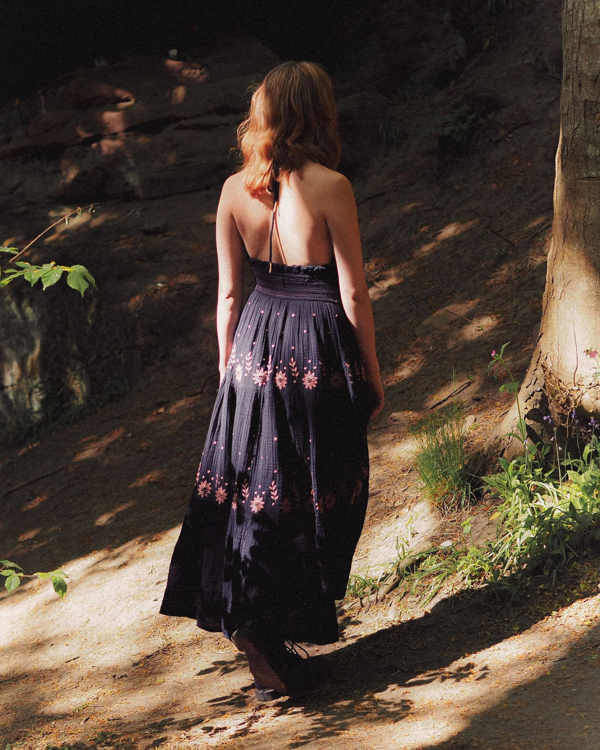 A woman with long, wavy ginger hair wearing a black dress with pink floral embroidery, walking on a dirt path in a wooded area with sunlight filtering through the trees.