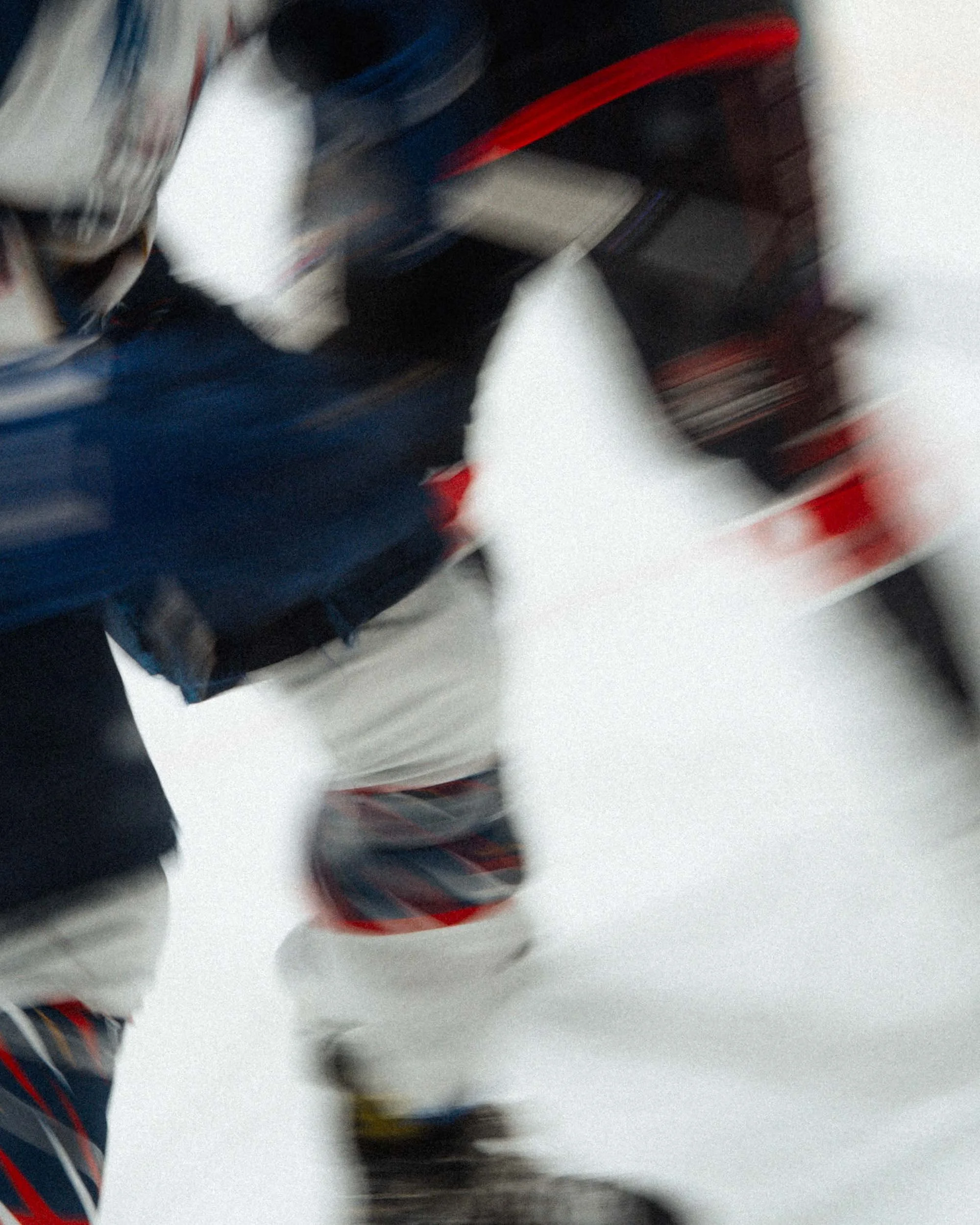 Blurred image of a person wearing a navy blue sports jersey and white hockey pants, holding a hockey stick, on an ice rink.