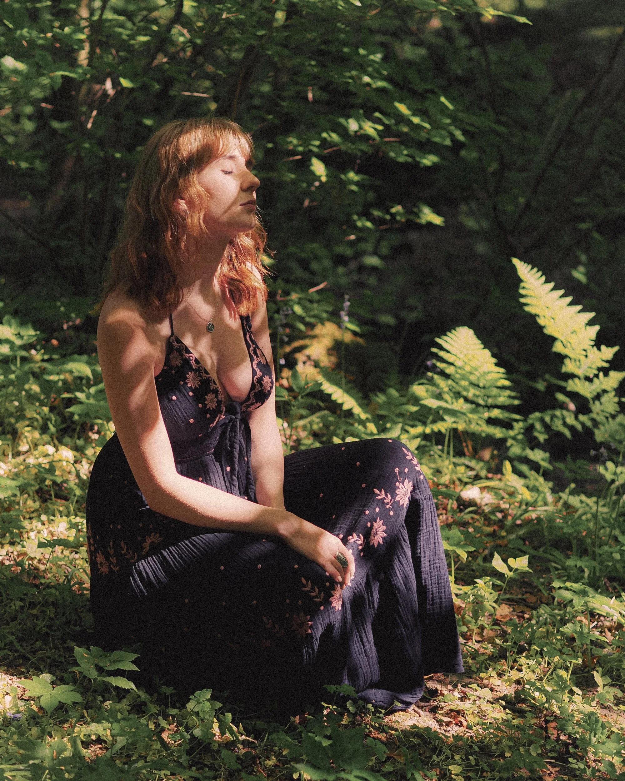 A woman with ginger hair and a floral dress sitting in a lush, green forest, with eyes closed and a peaceful expression.