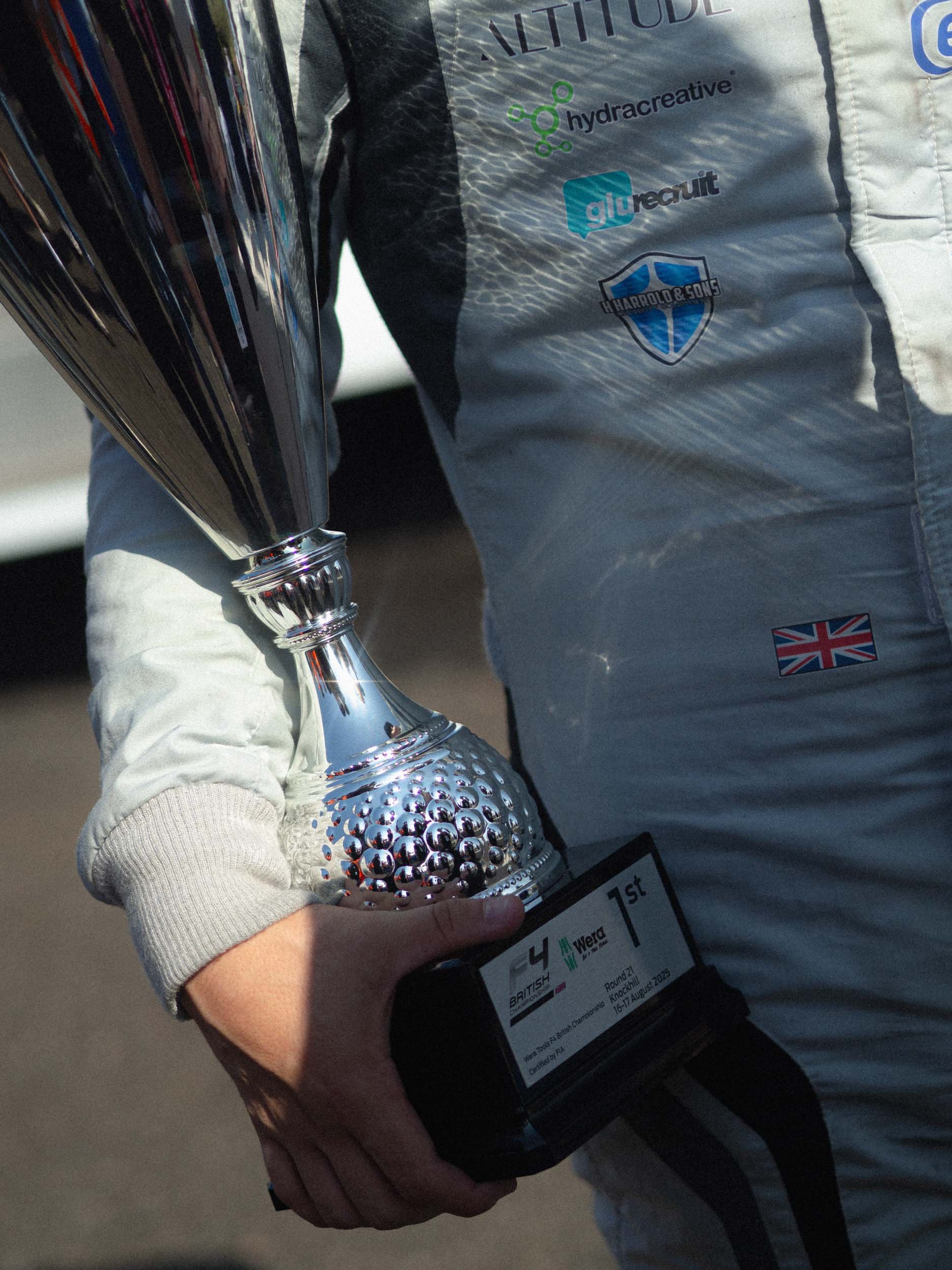 Person in racing suit holding a silver trophy and a small plaque indicating first place at the F4 British Championship.