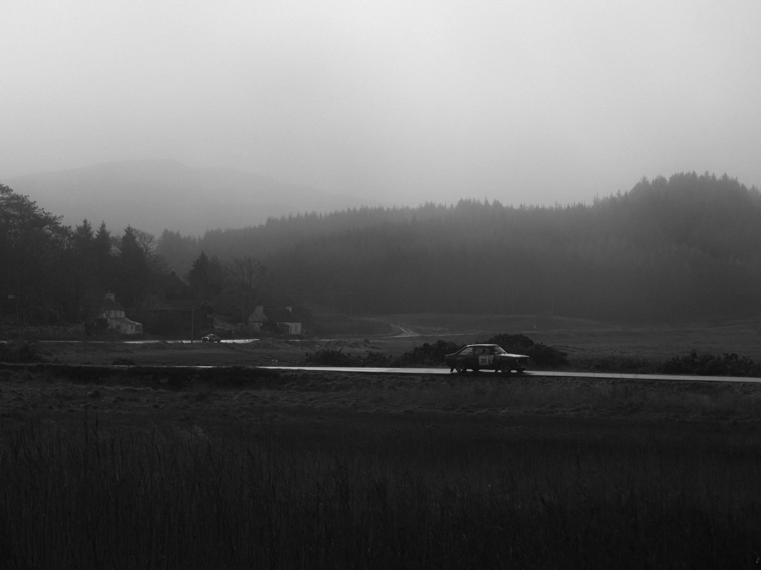Black and white landscape scene with a rally car driving on a road. Houses, trees, and mountains in the background. A dense fog rolls over the distant mountains.
