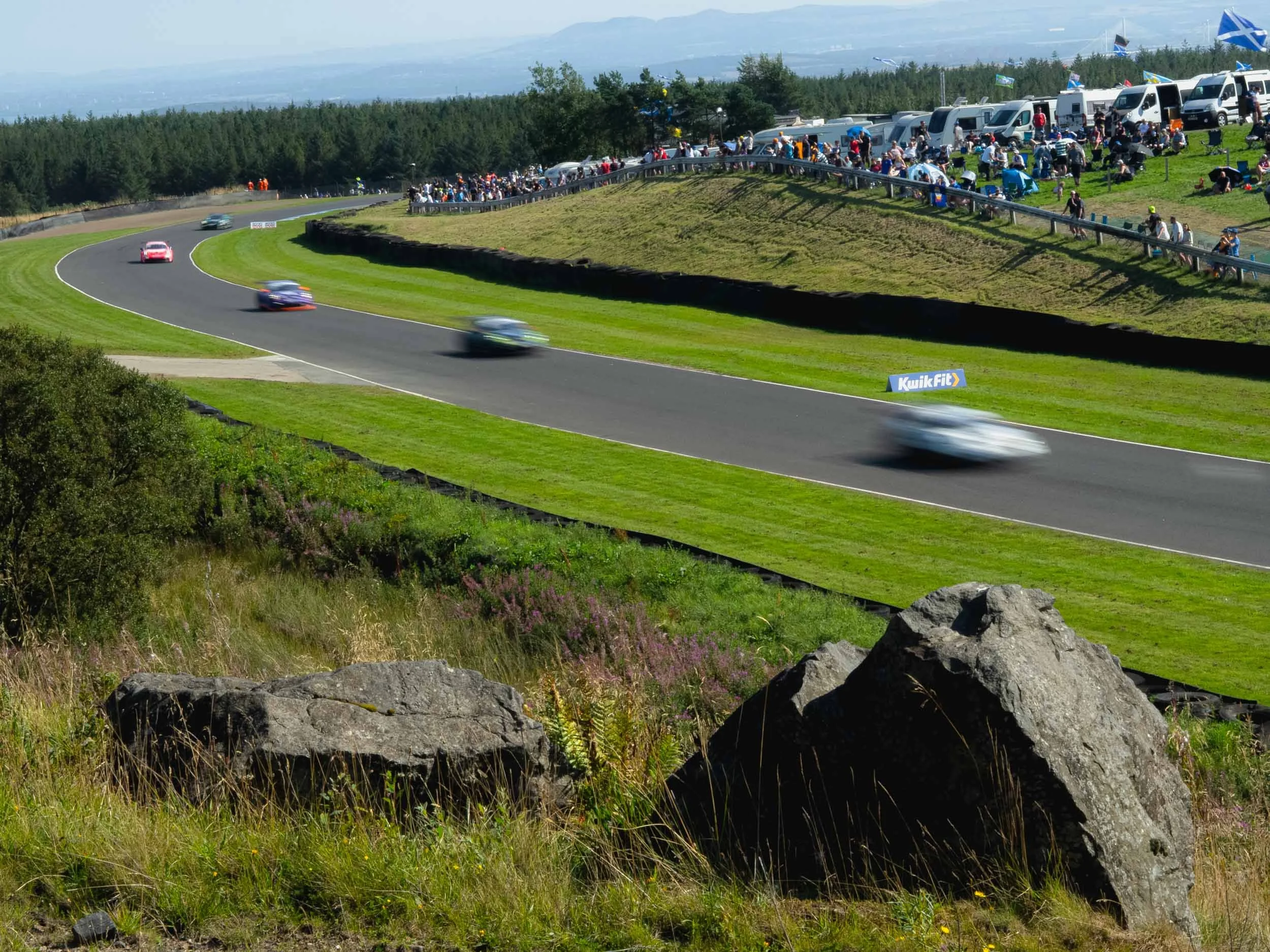 A race track with several cars racing on a sunny day, spectators watching from the grassy area and parked cars in the background.