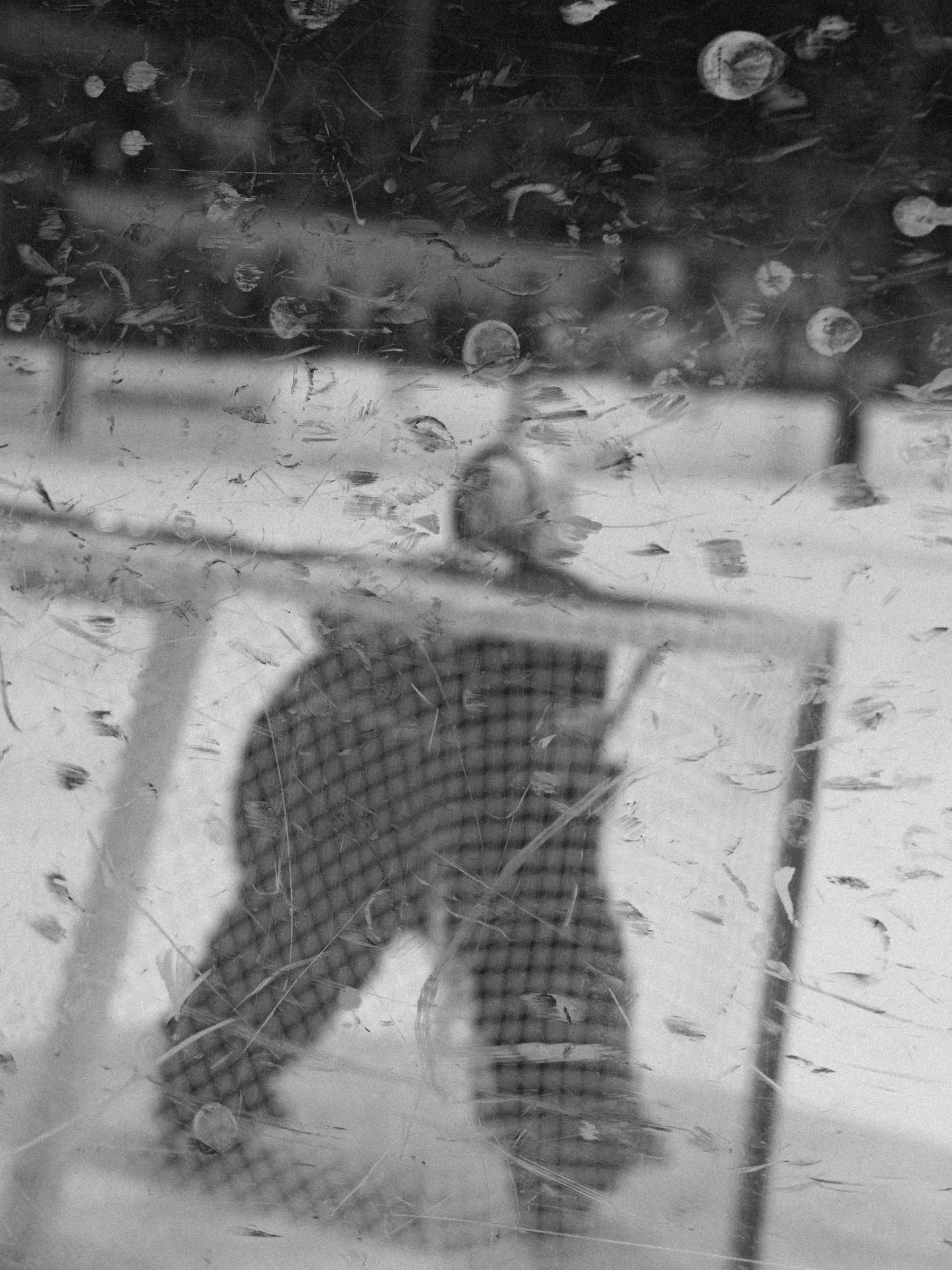 A goalie viewed through a dirty glass surface, with dents and scratches obscuring the view. The person appears to be wearing a hockey jersey and standing in front of the goal net.