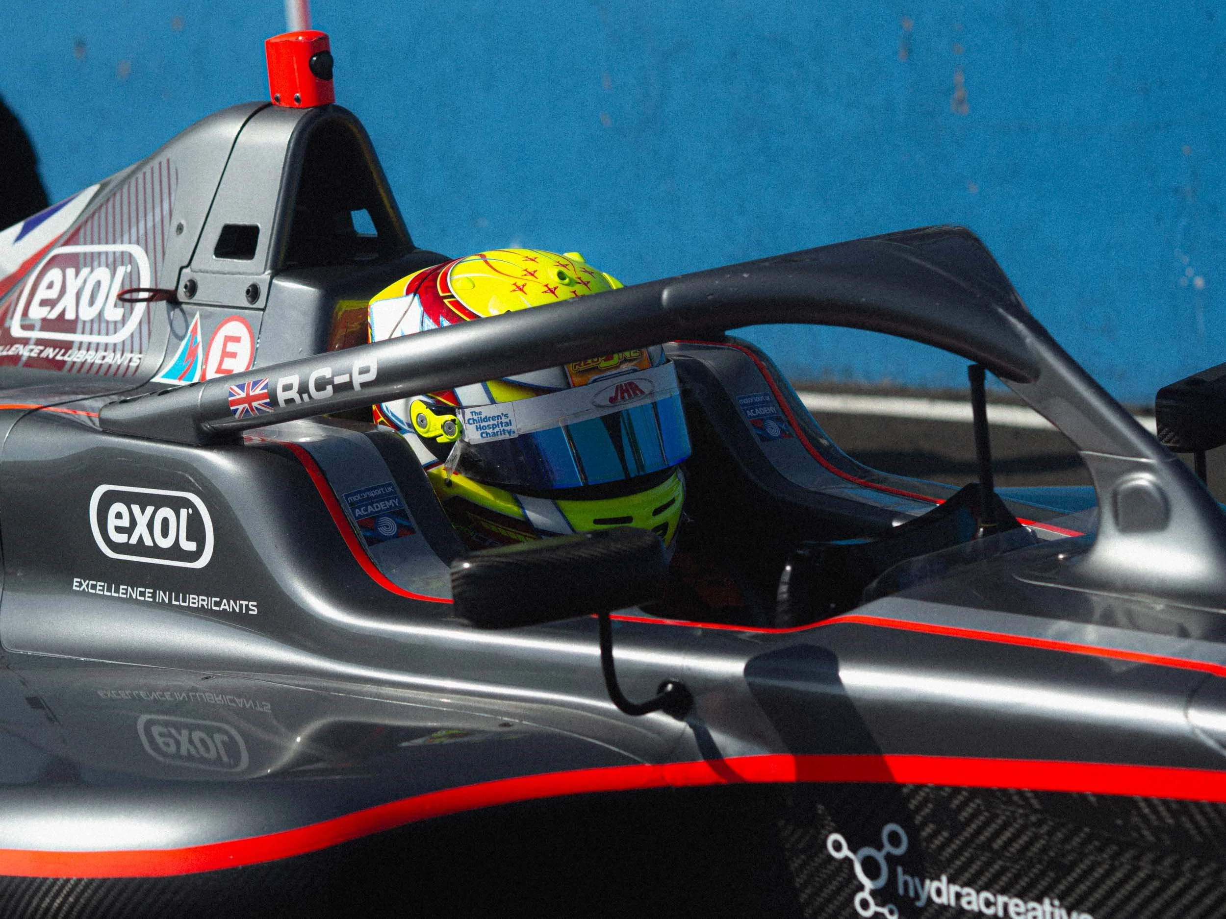 Close-up of a race car with a driver inside wearing a helmet. The car has various sponsor logos like 'exol' and is parked against a blue background.