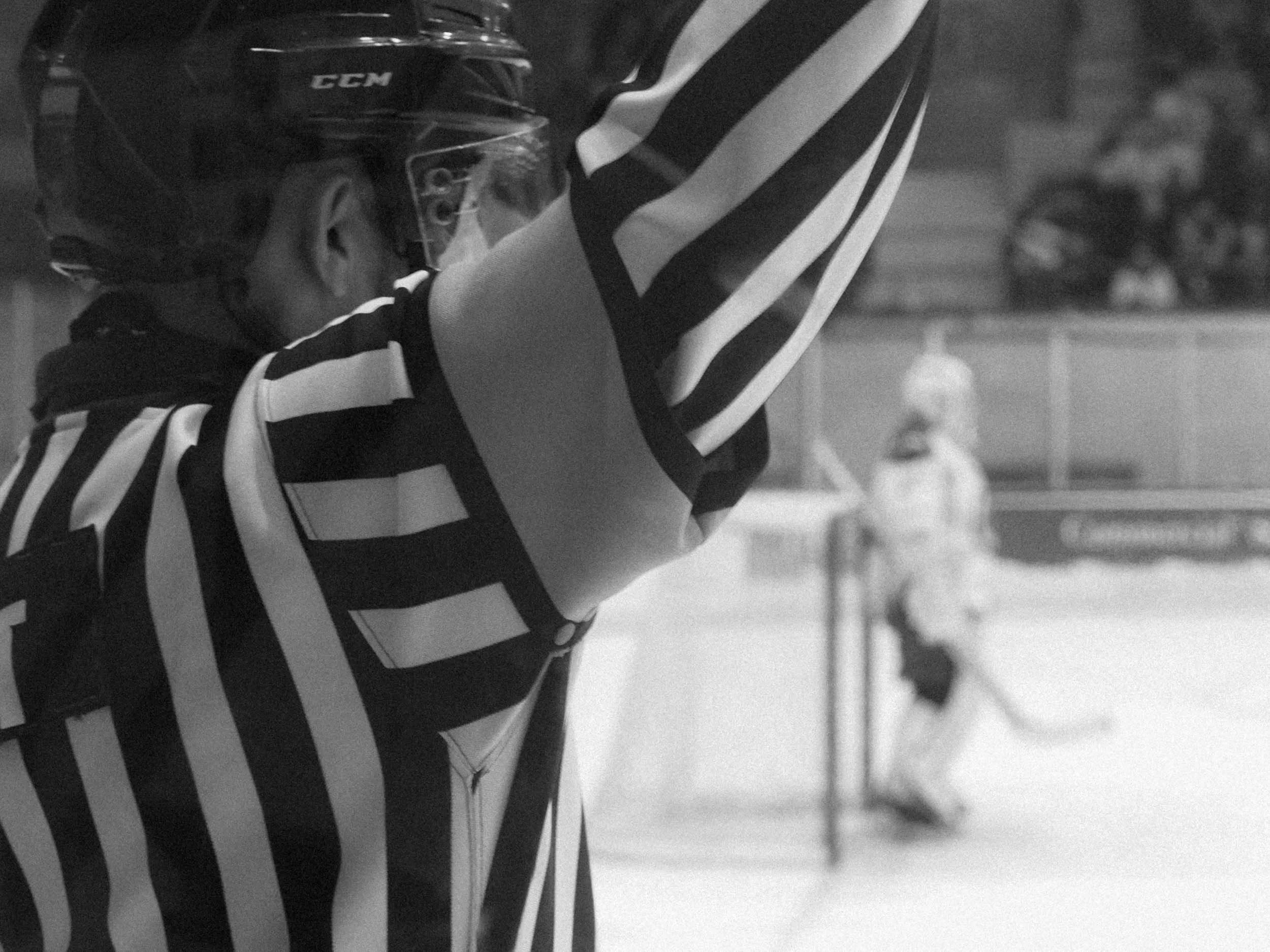 Hockey referee in black and white striped shirt raising hand, with a hockey player in the background on the ice rink.