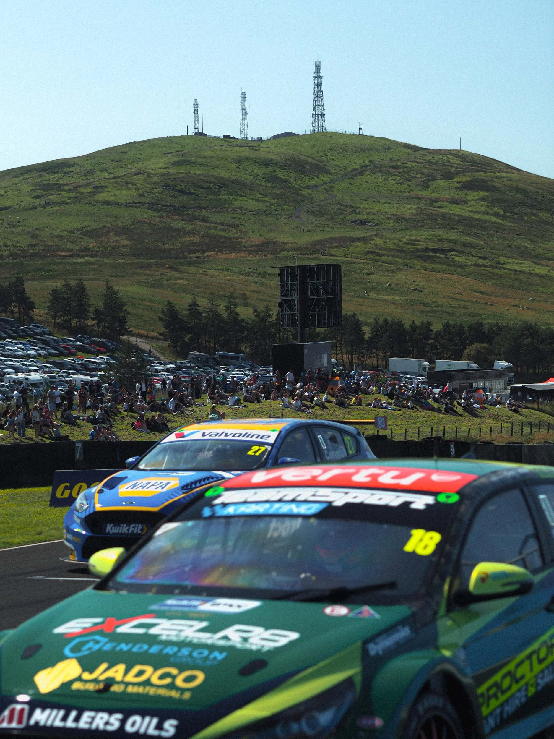 Race cars on a track, with spectators watching from a hillside. In the background, a mountain with communication towers.
