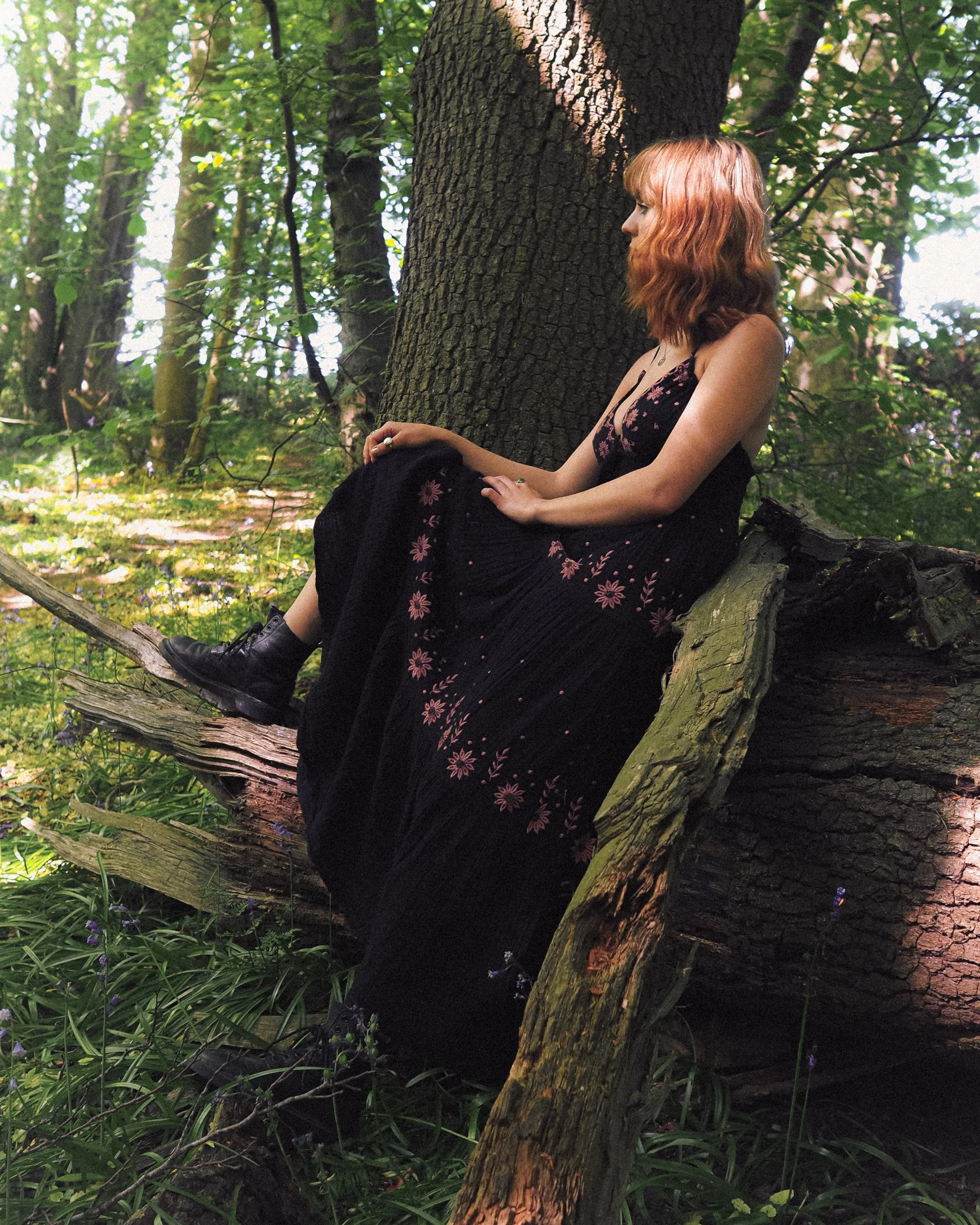 A woman with ginger hair sitting on a fallen tree in a lush forest, wearing a black dress with floral embroidery and black boots.