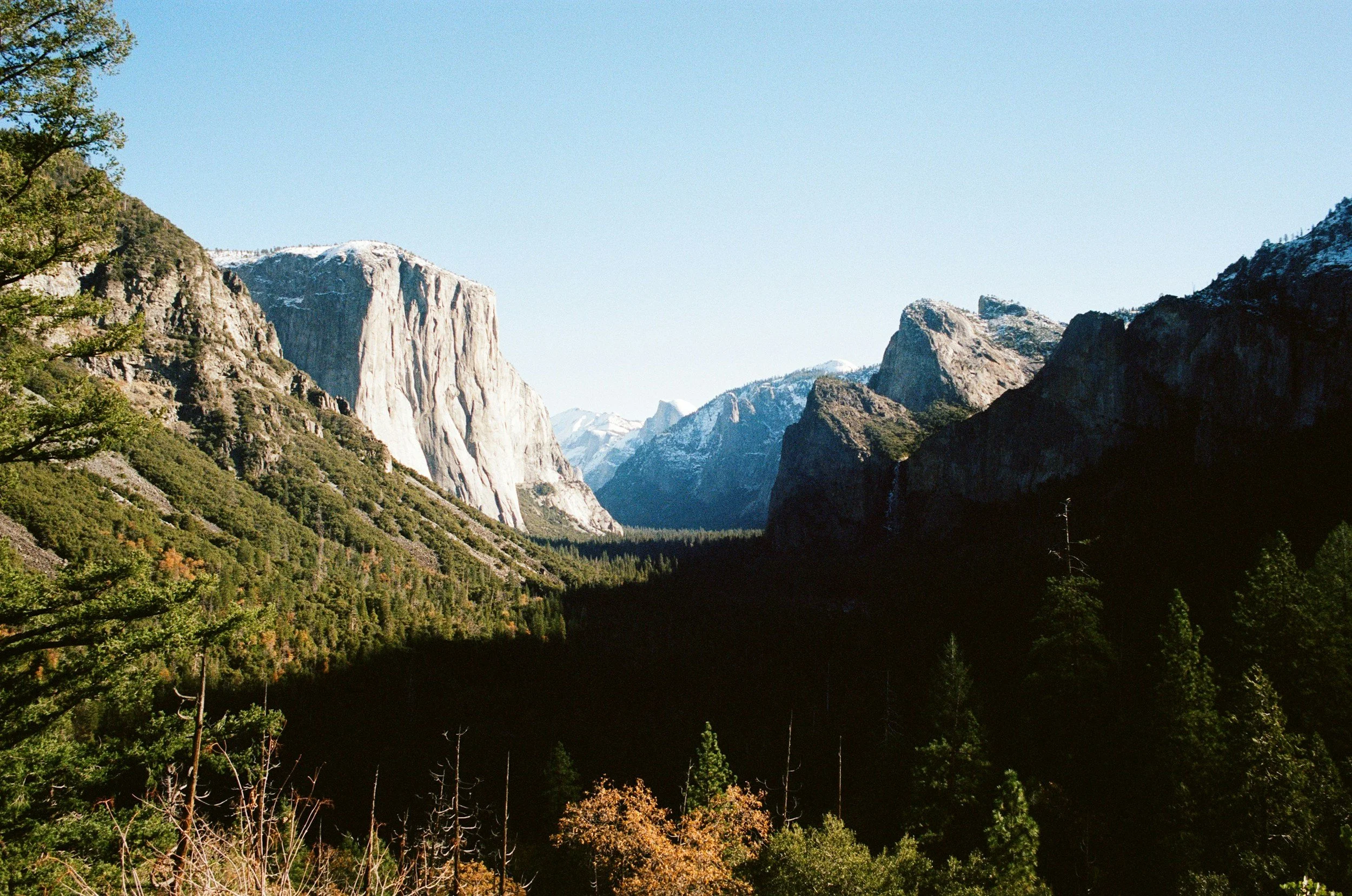 Scenic view of a mountain valley with large granite cliffs, dense green forest, and snow-capped peaks under a clear blue sky.