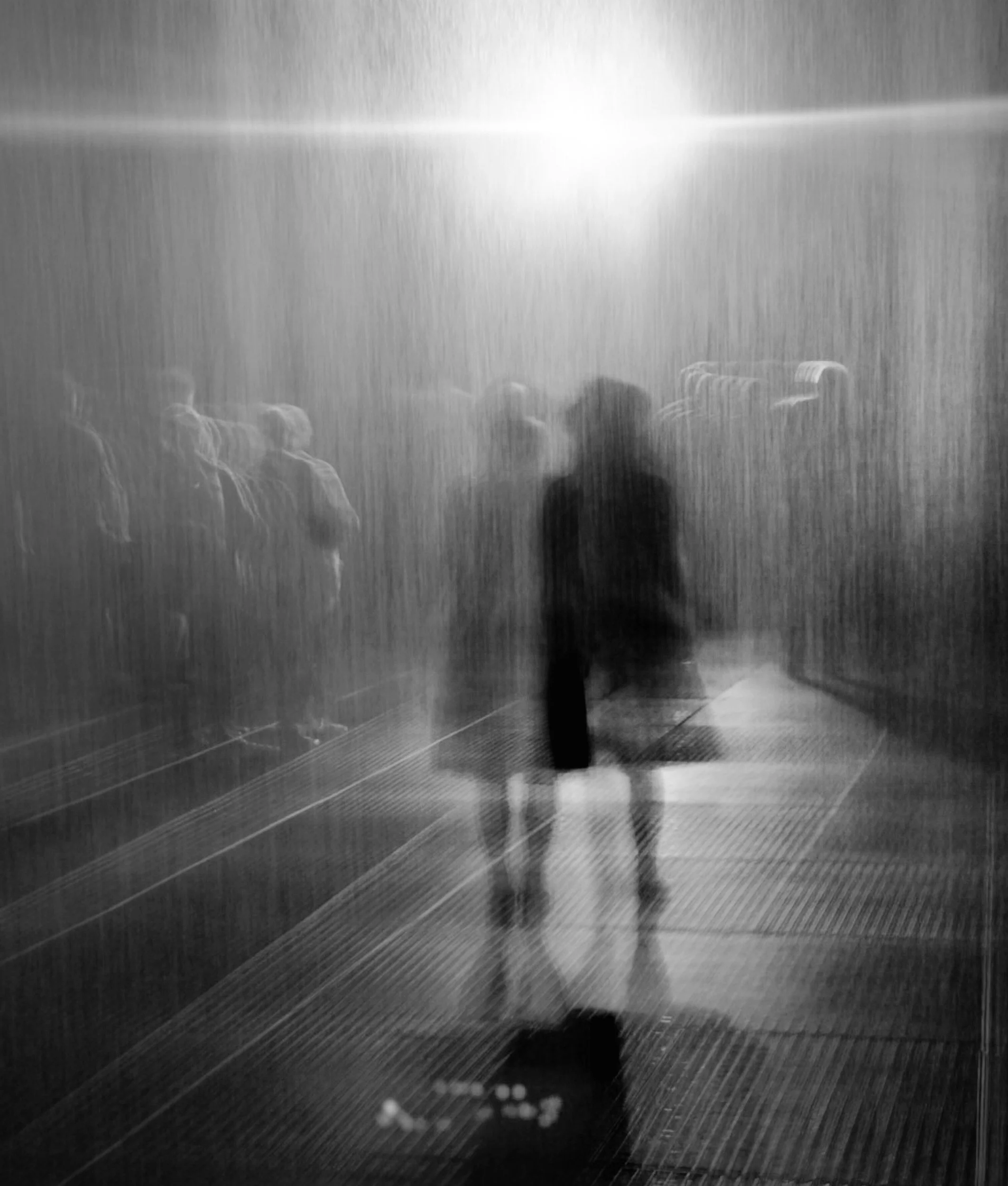 Blurry black and white image of a person standing in front of a rain-covered glass wall or window, with other people visible behind the glass.