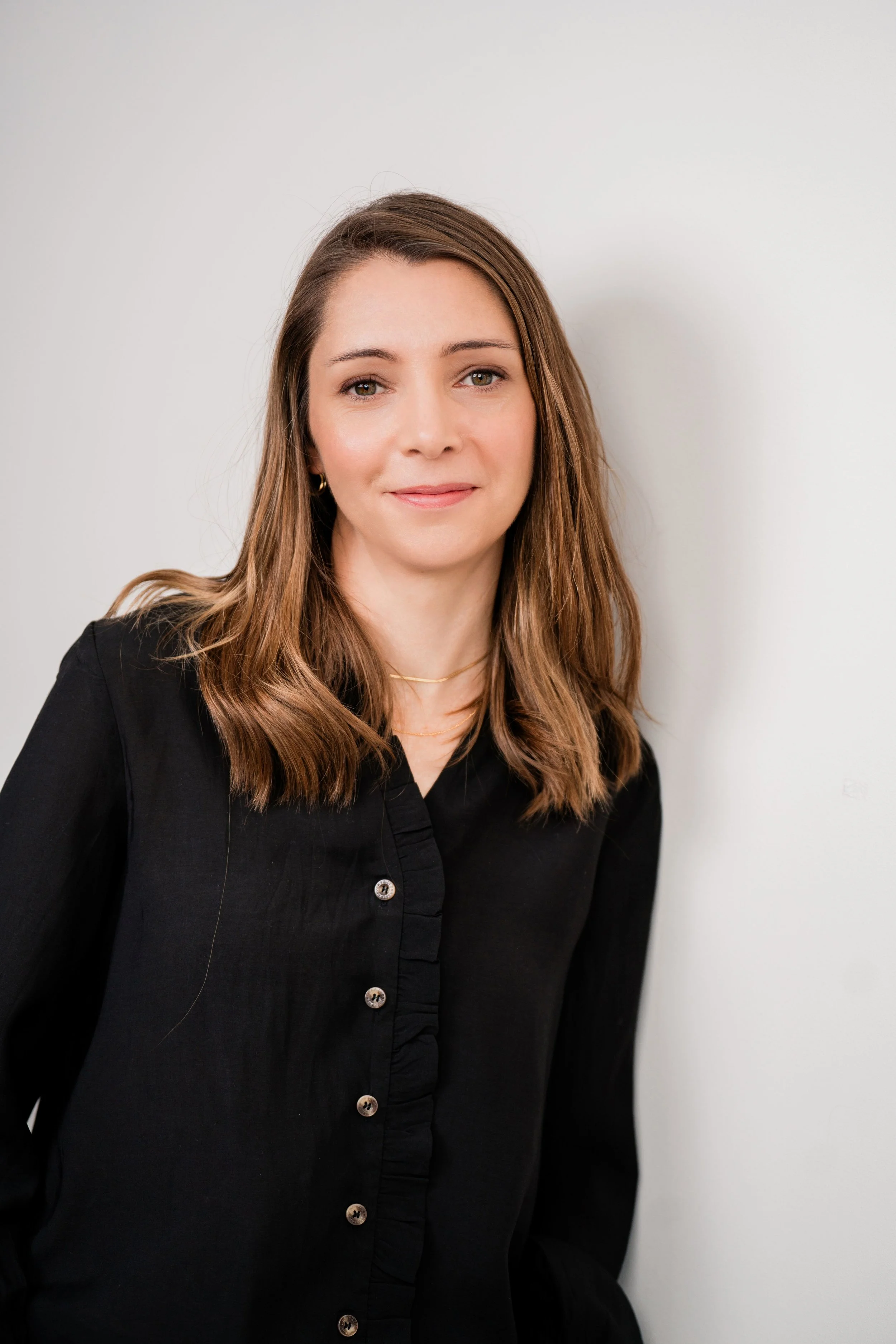 A woman with shoulder-length light brown hair stands against a plain white background. She is wearing a black button-up shirt with ruffled detailing. Her expression is calm and composted, and the image is professionally lit and styled.